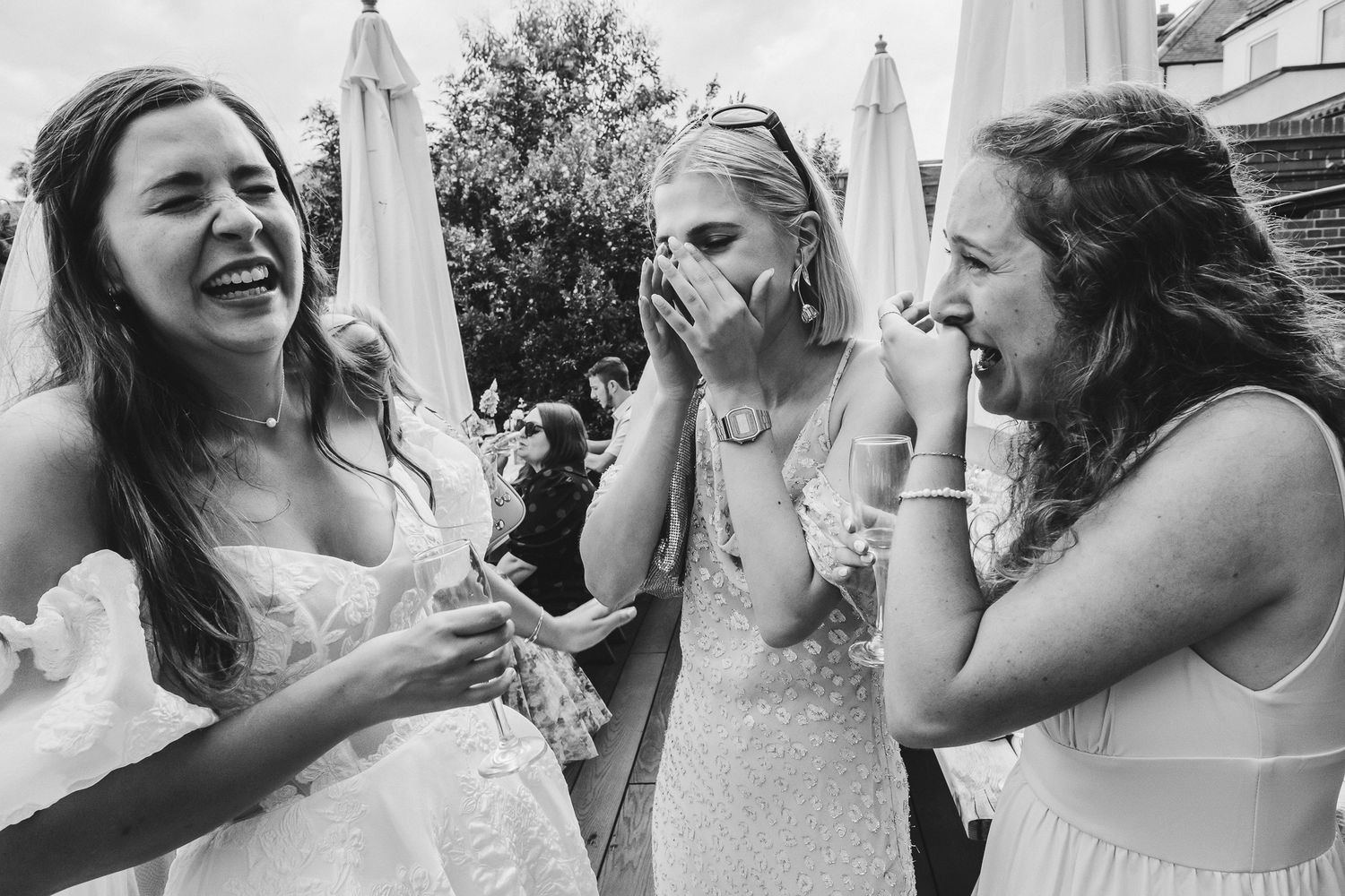 Black and white candid photo of three people in white dresses laughing together outdoors at a celebration.