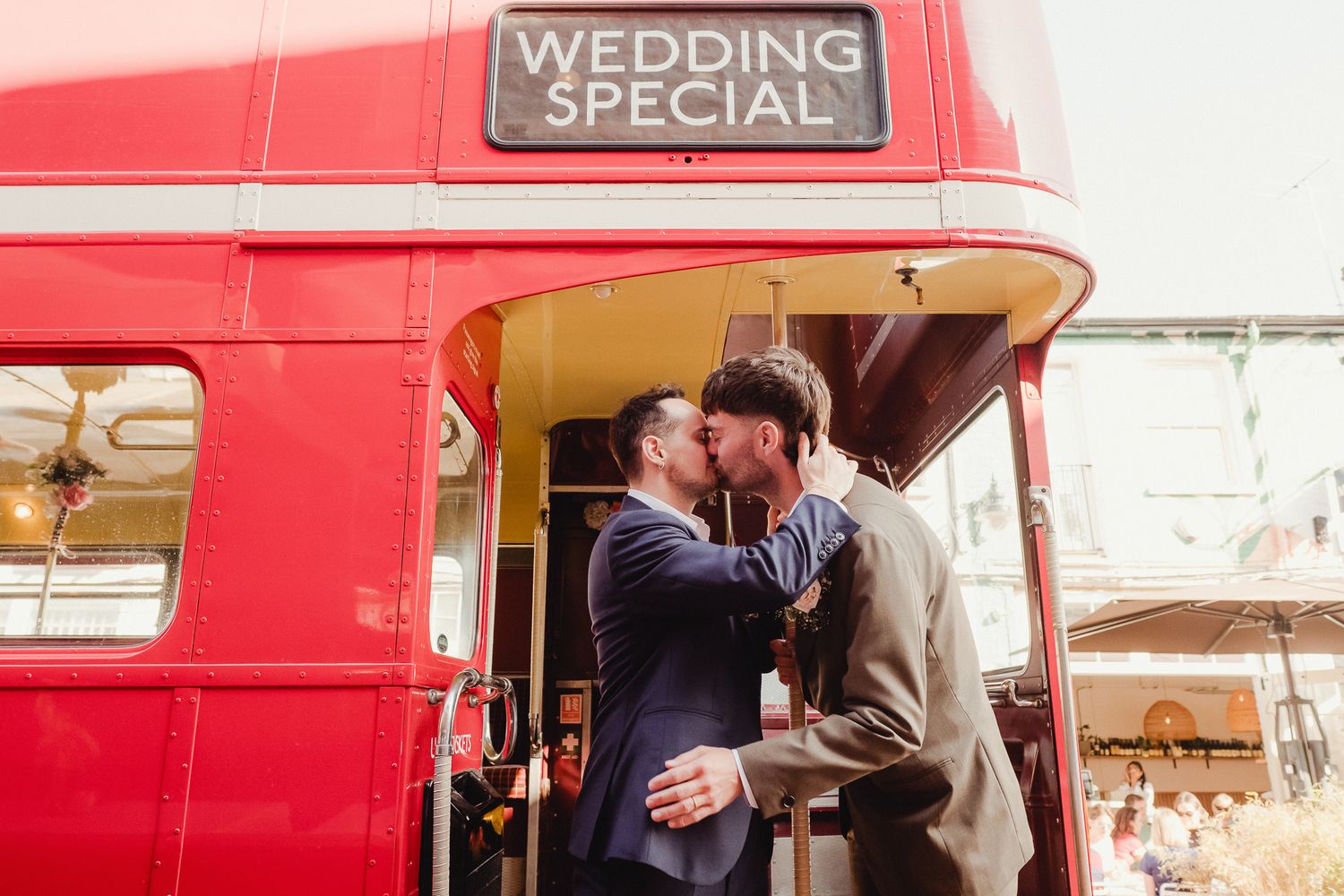 A couple embraces and kisses next to a vintage red double decker bus during their wedding celebration.