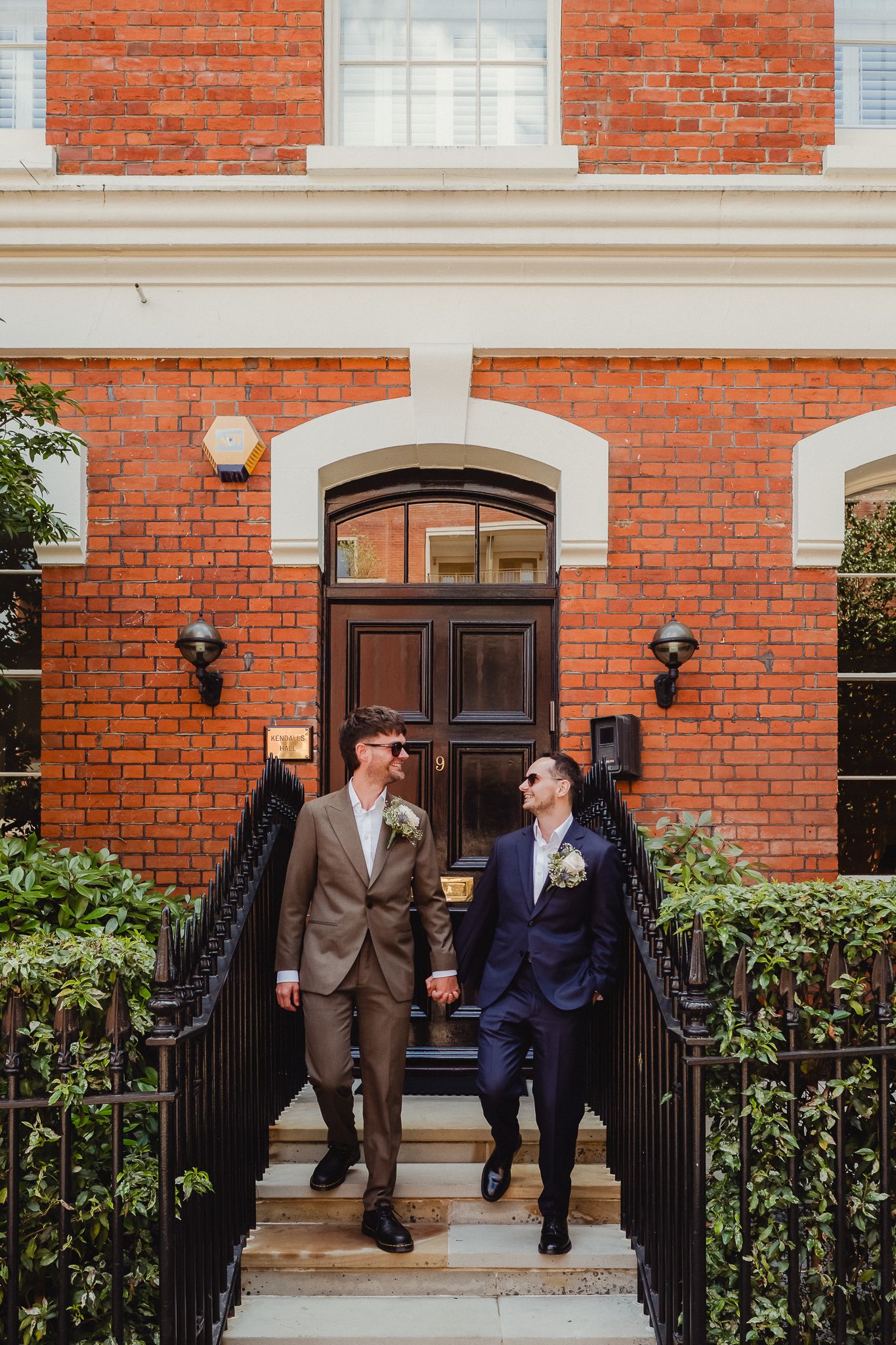 Three people in formal attire share a laugh together outside a red brick building with white trim and black railings.
