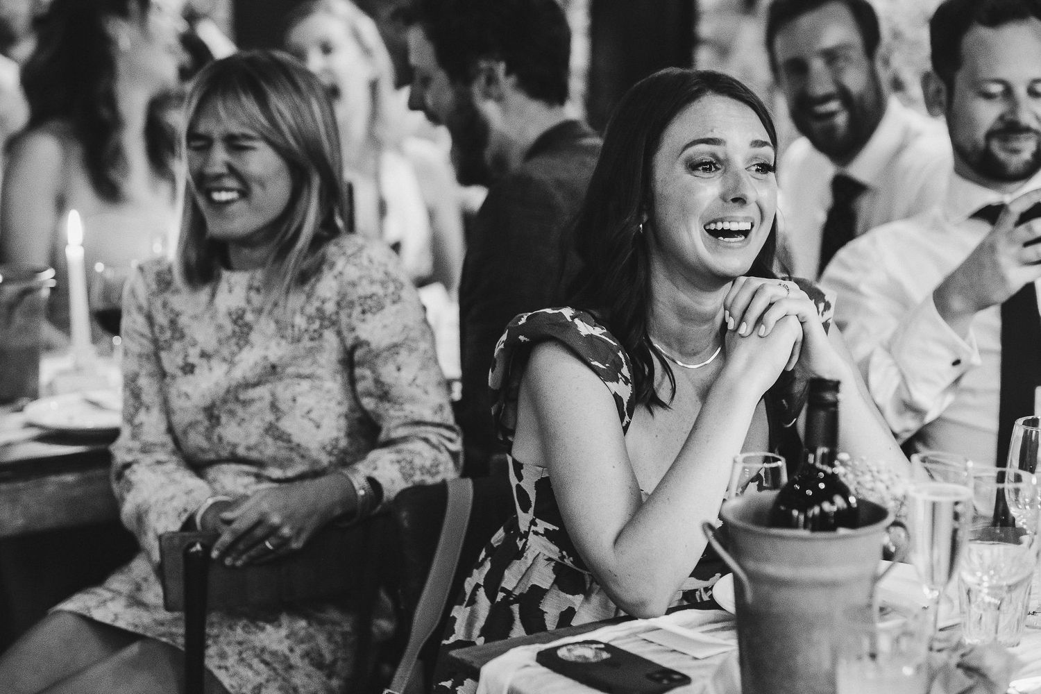Wedding reception guests laugh and smile while seated at tables during a celebration.