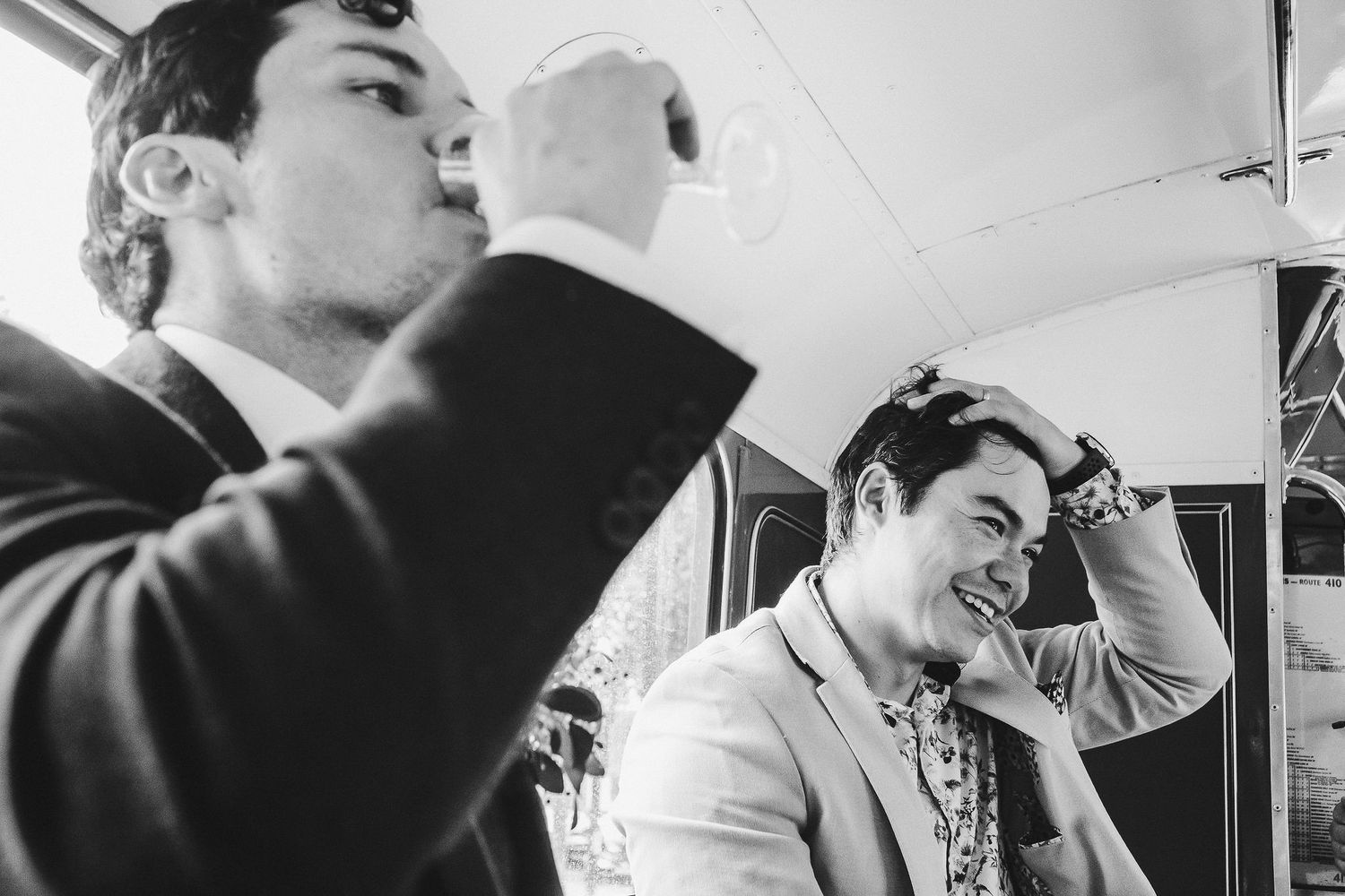 Two men in formal attire sharing a lighthearted moment while drinking inside a vehicle.