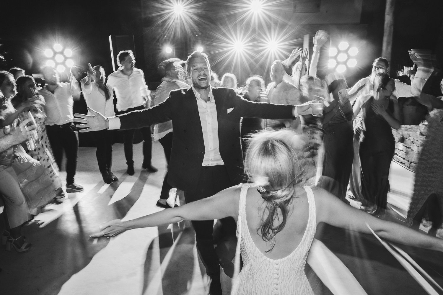 Wedding guests dance and celebrate on a crowded dance floor with bright overhead lighting creating a festive atmosphere.
