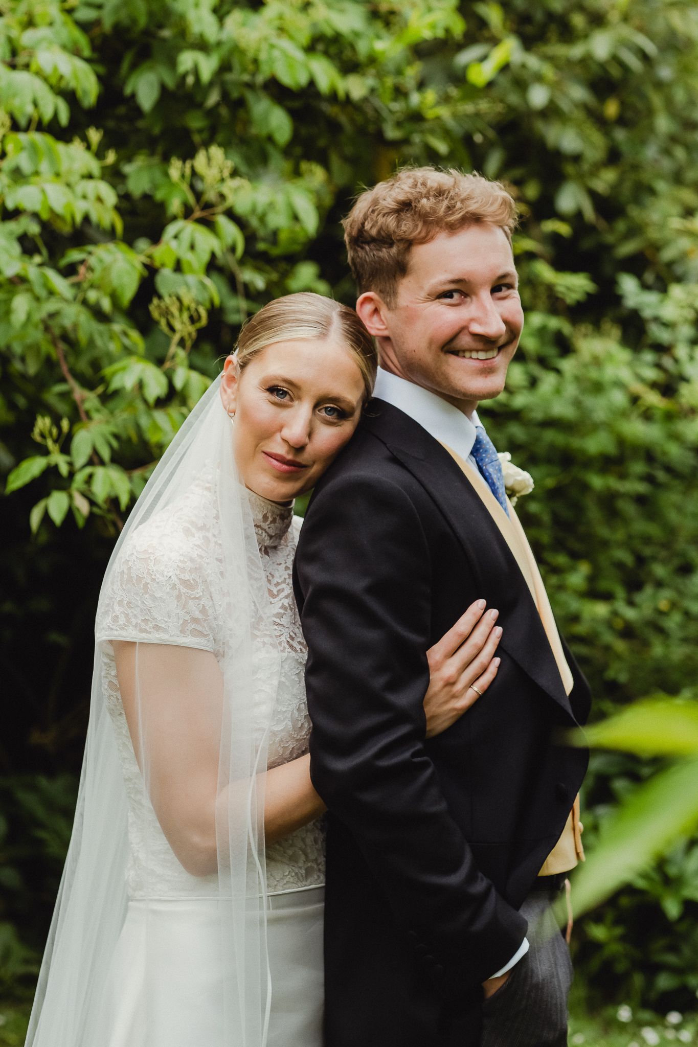 A newly married couple shares a playful moment outdoors against a backdrop of lush green foliage.