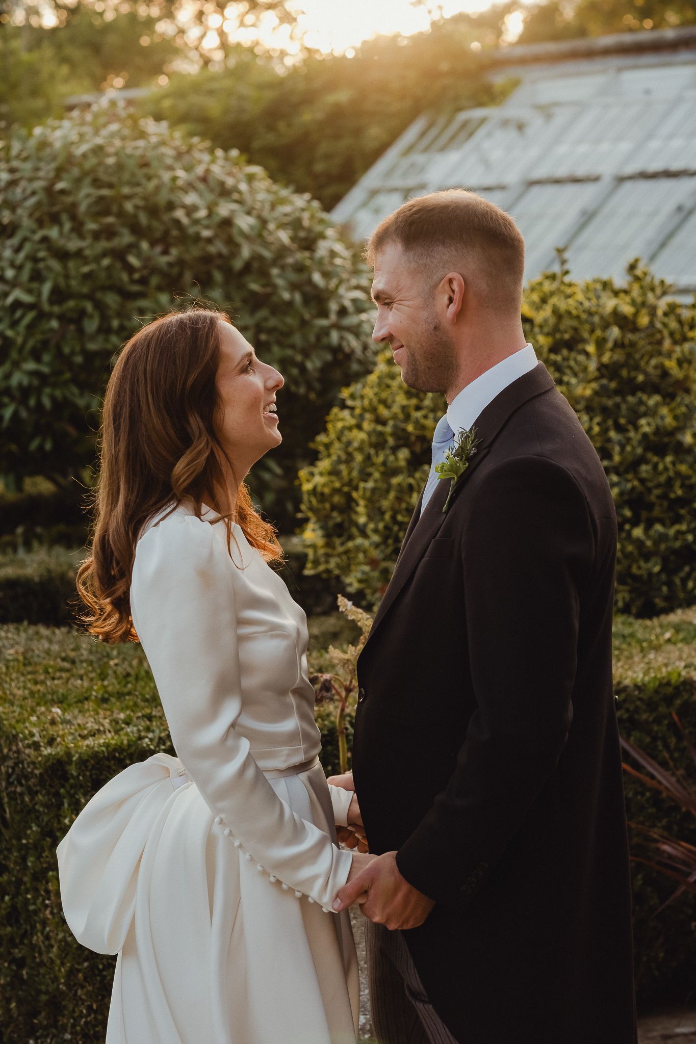 A couple shares an intimate moment in a garden setting during sunset, with the bride wearing an elegant white dress.