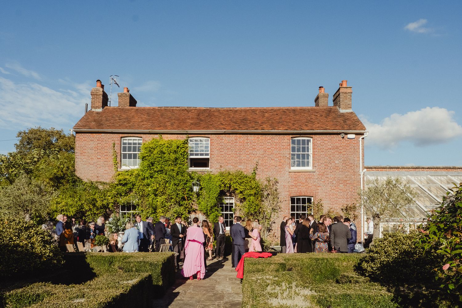Row of wedding guests gather outside a rustic red brick country house on a sunny summer day.