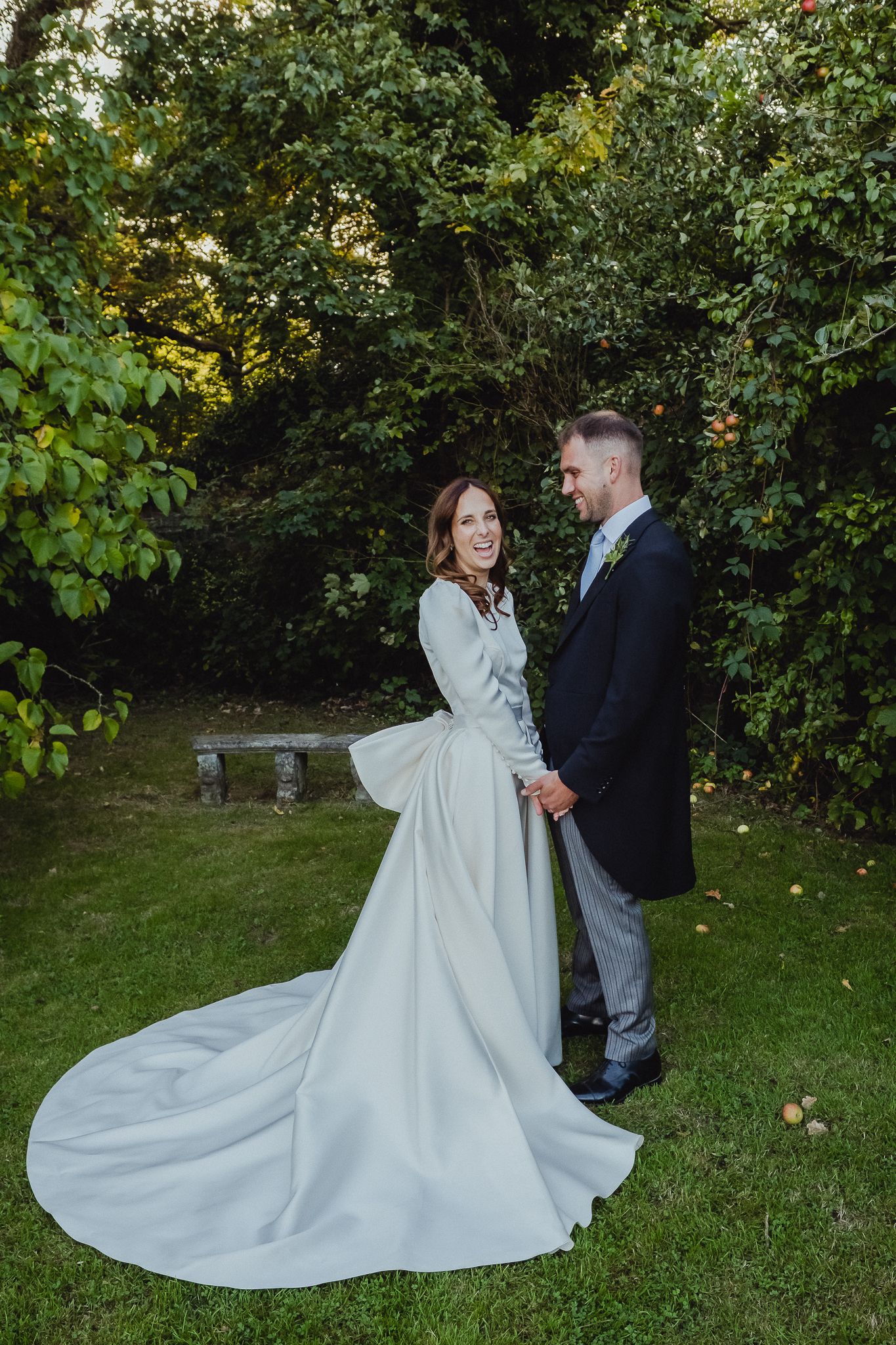 Bride in flowing white dress with long train poses with groom in outdoor garden setting during wedding photoshoot.