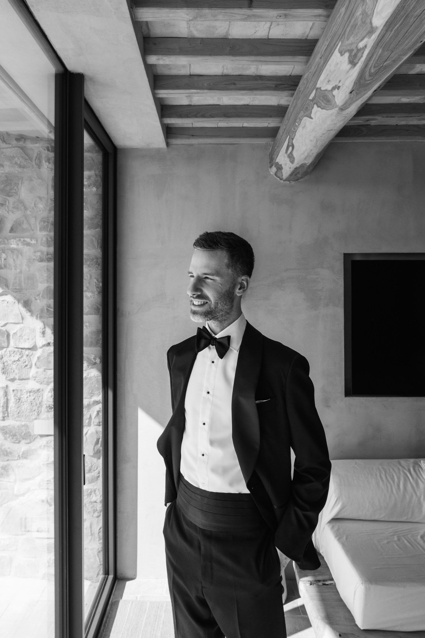Black and white portrait of a groom in a tuxedo and bow tie standing by a large window in a rustic room with exposed wooden beams.