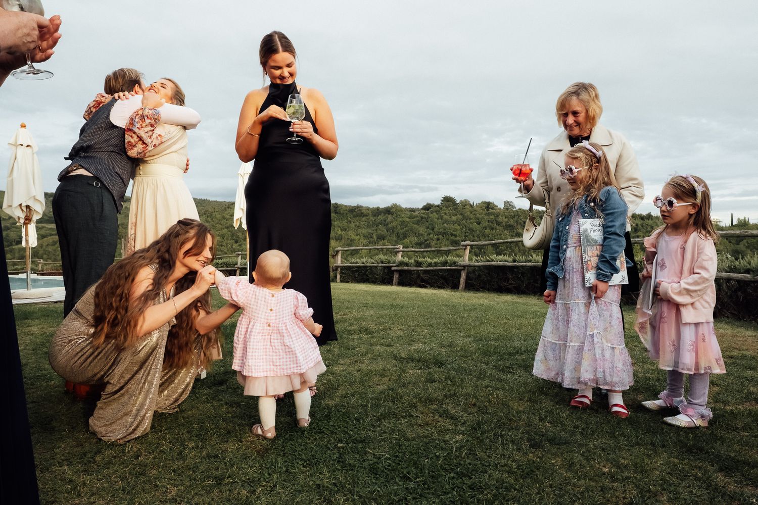 Toddler in a pink dress takes first steps on a grassy lawn at an outdoor party while family and young girls look on and smile.