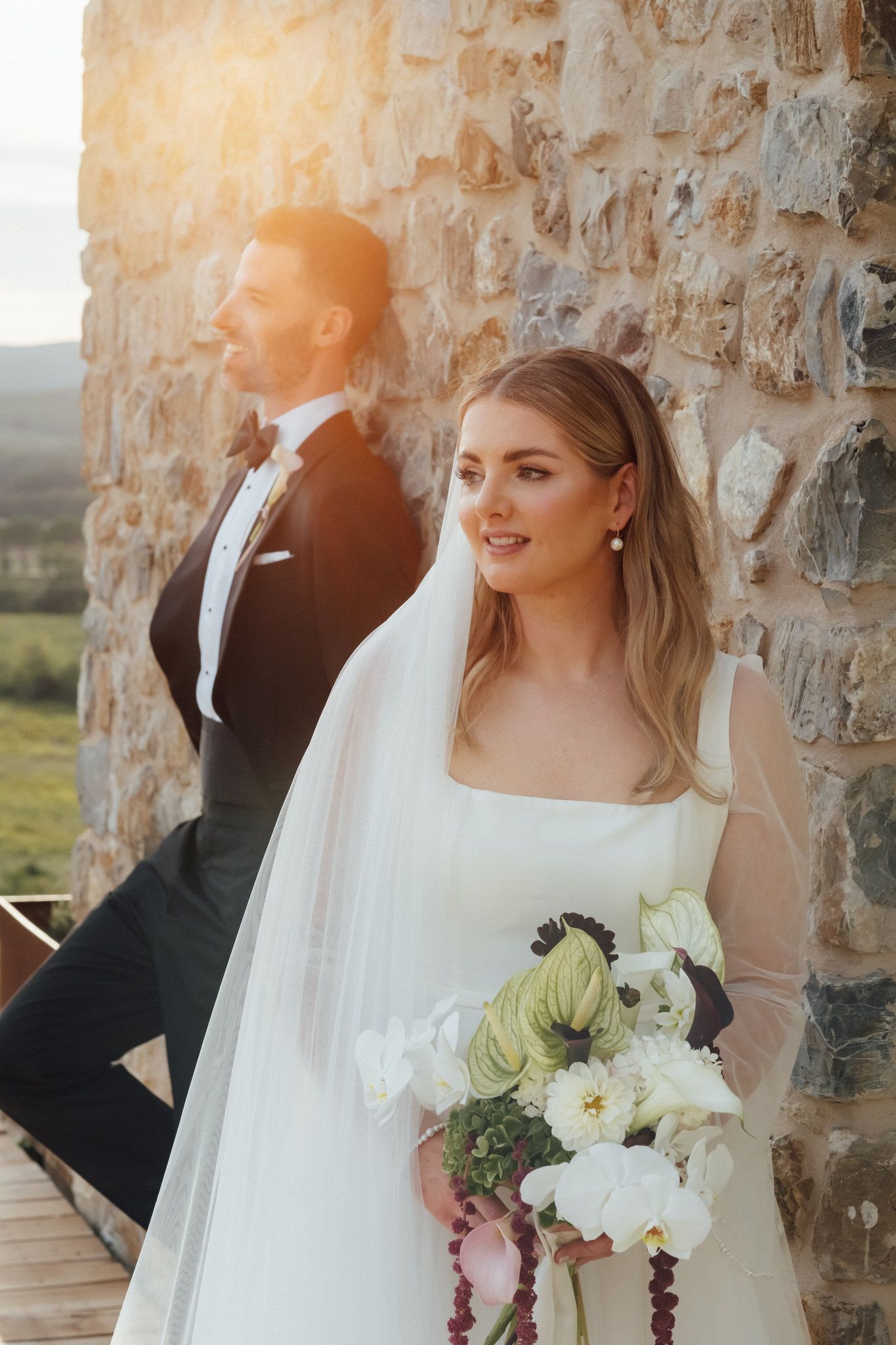 Bride in a white gown and veil holds a lush bouquet beside a groom in a black tuxedo against a rustic stone wall at golden hour.