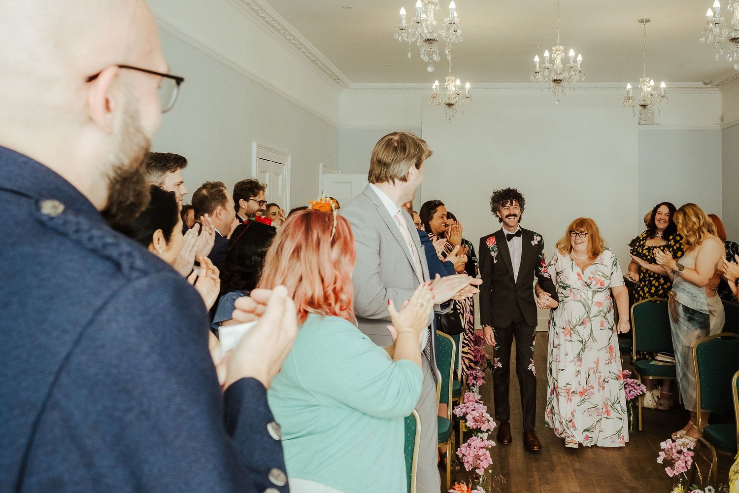 Wedding guests applaud and smile as a bride and groom walk down the aisle of a bright venue lined with pink flowers and chandeliers.