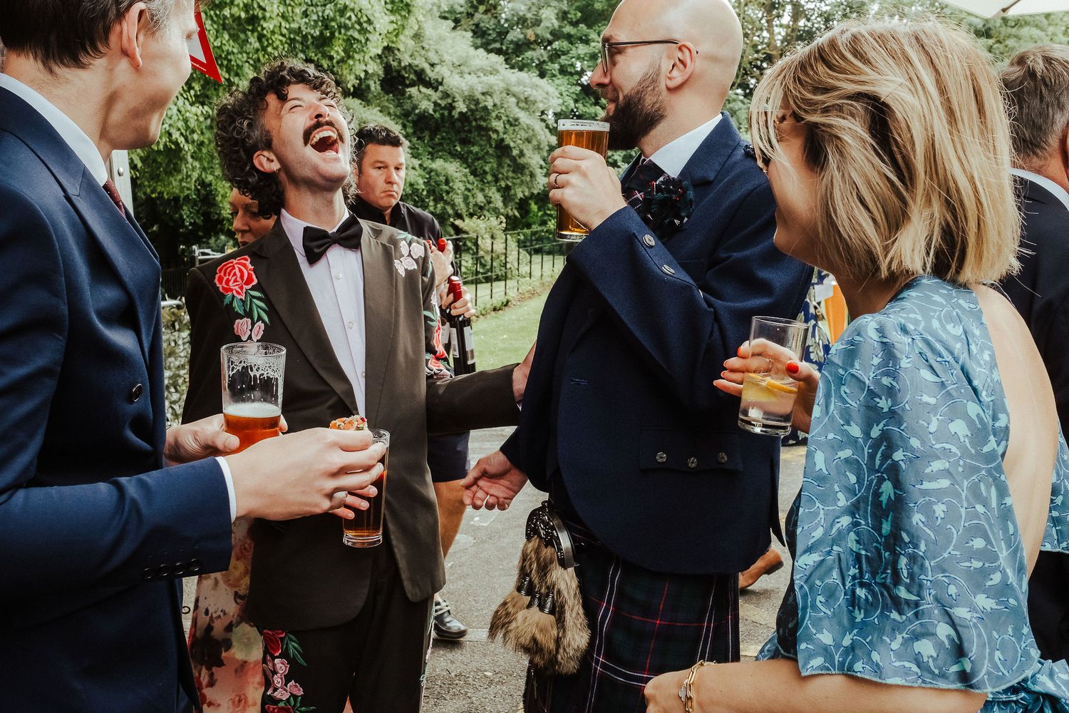 Wedding guests in formal attire laugh and raise beer glasses while chatting outdoors, including a man in a bow tie and a kilt.