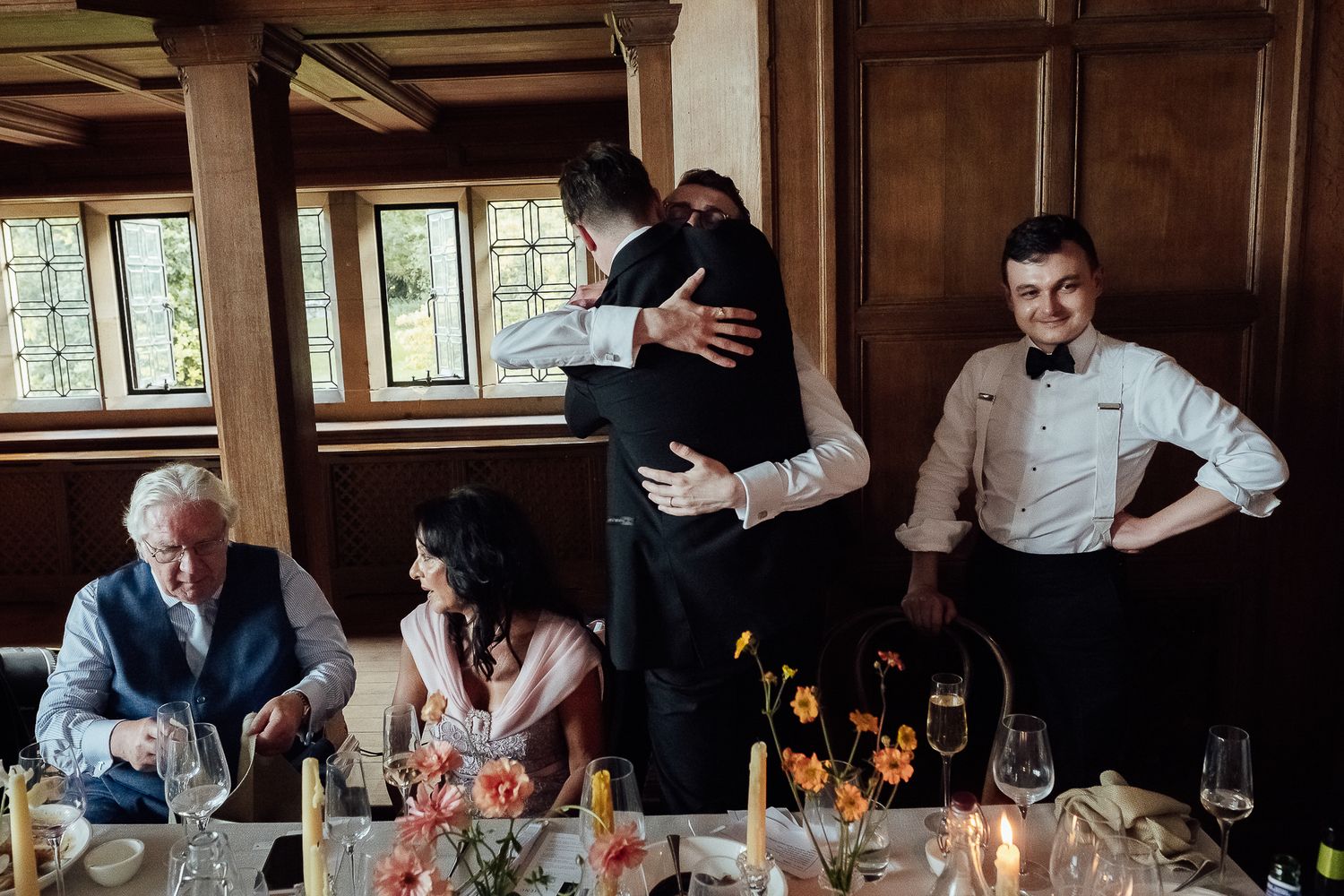 Two men embrace at a wedding reception dinner table while guests watch in an elegant wood-paneled dining room.