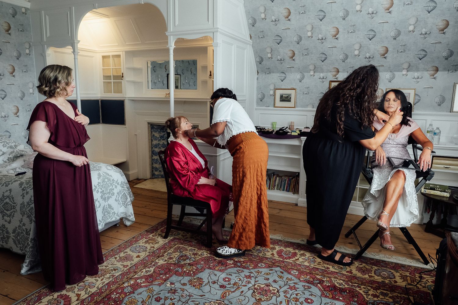 Bridesmaids getting hair and makeup done in an elegant room with floral wallpaper before a wedding ceremony.