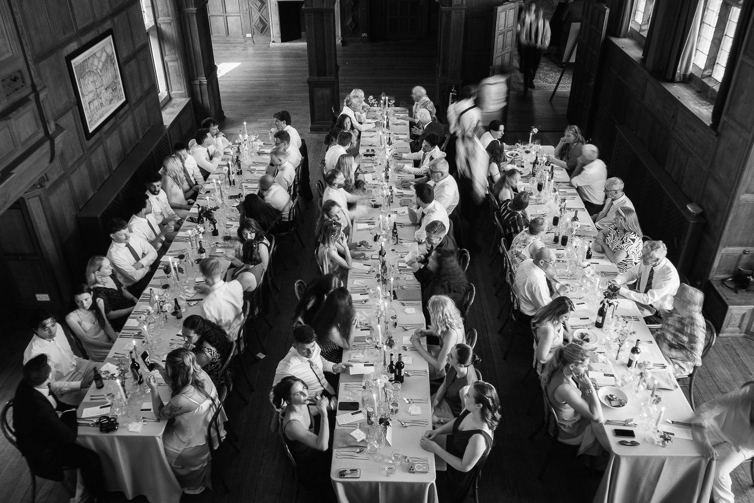 Black and white aerial view of guests dining at long banquet tables in an elegant wood-paneled hall.