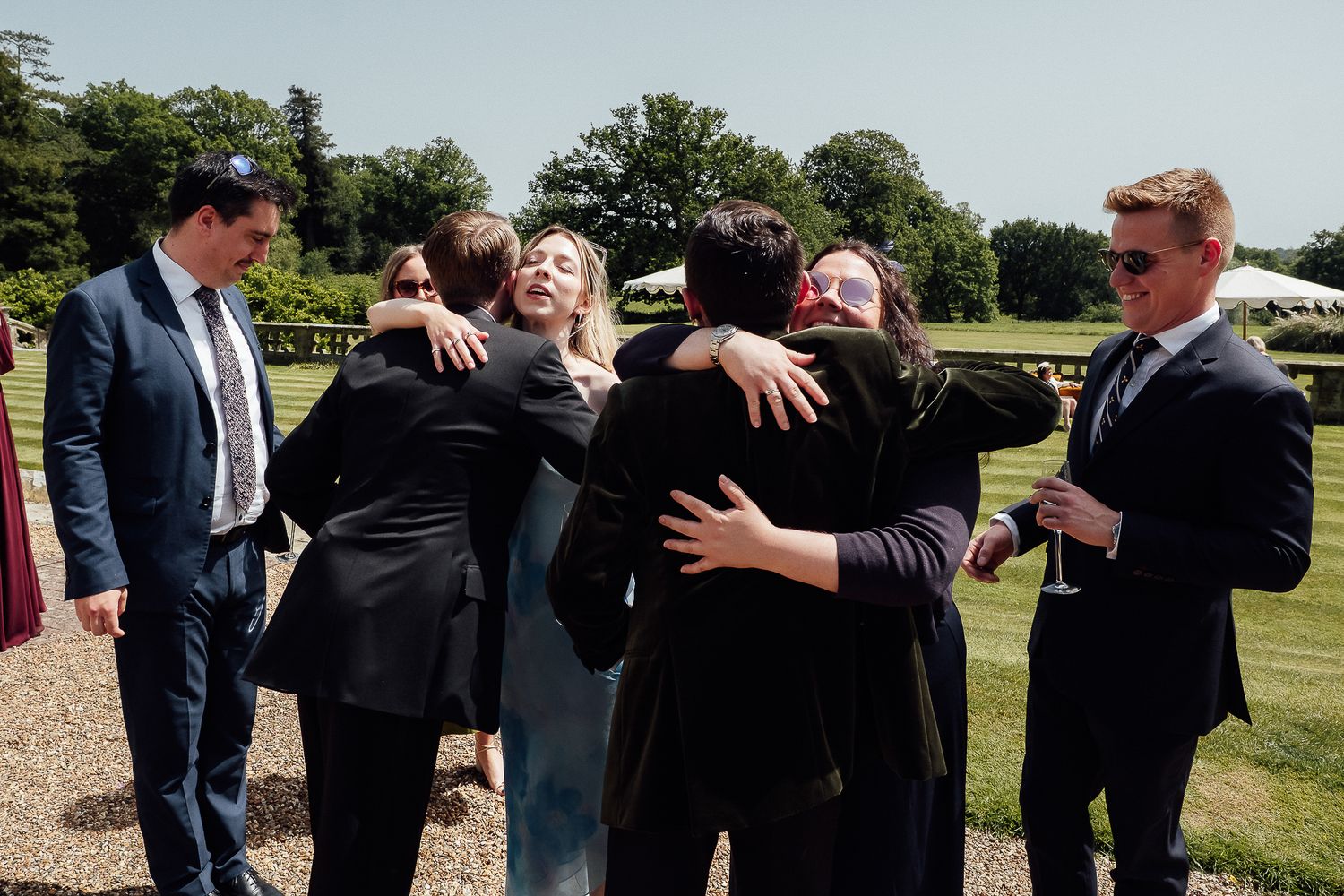 Group of elegantly dressed guests sharing warm hugs at an outdoor wedding reception on a sunny day.