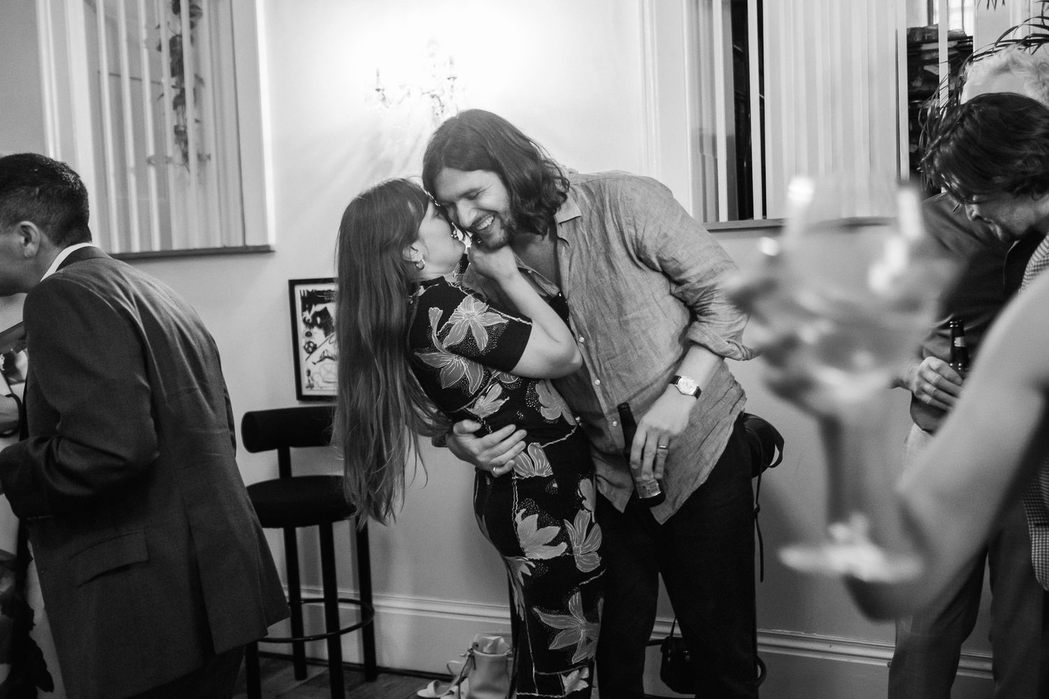 Black and white photo of a long-haired couple embracing and sharing an intimate moment at a lively indoor party with guests nearby.