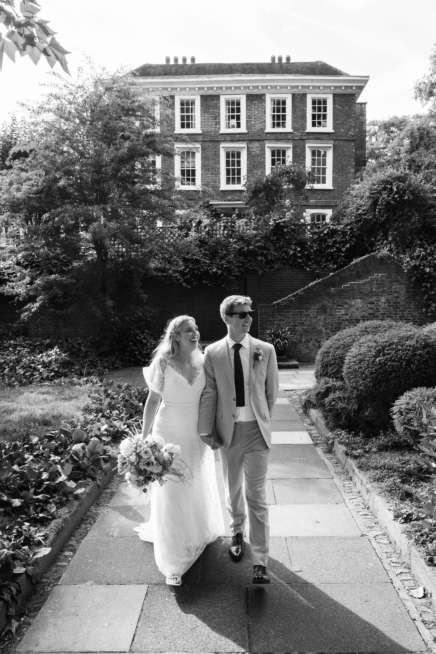 Bride in a flowing white gown holding a bouquet walks beside a groom in a light suit along a garden path toward a brick Georgian house.