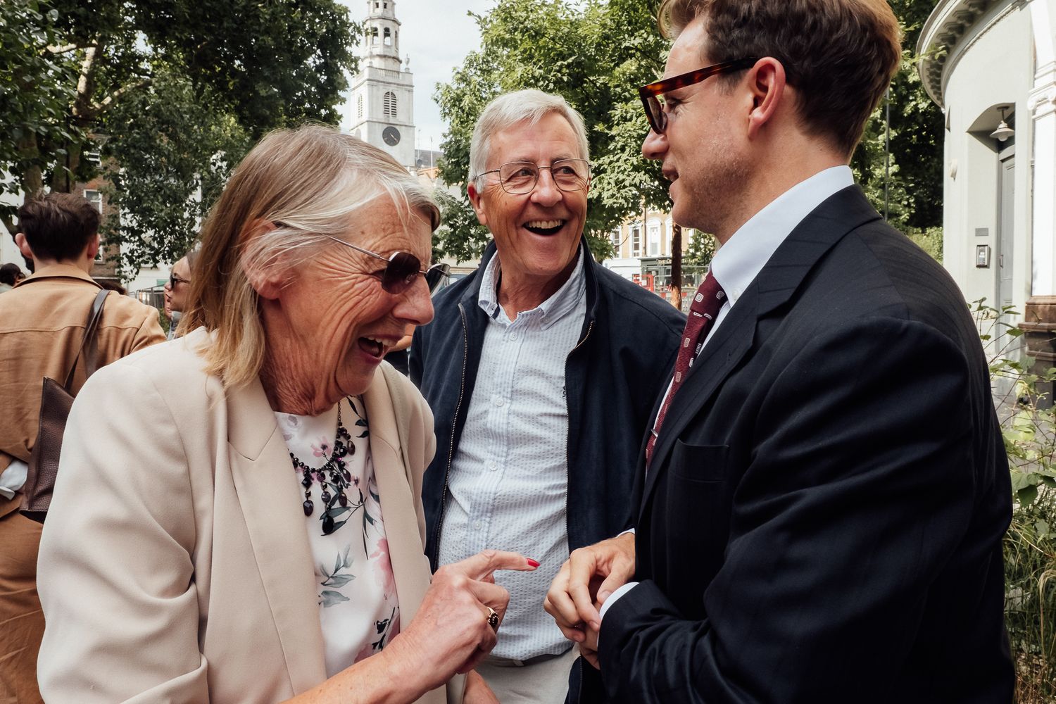 Three smiling people chat outdoors on a sunny day, an older woman laughing with a gray-haired man and a younger man in a suit.