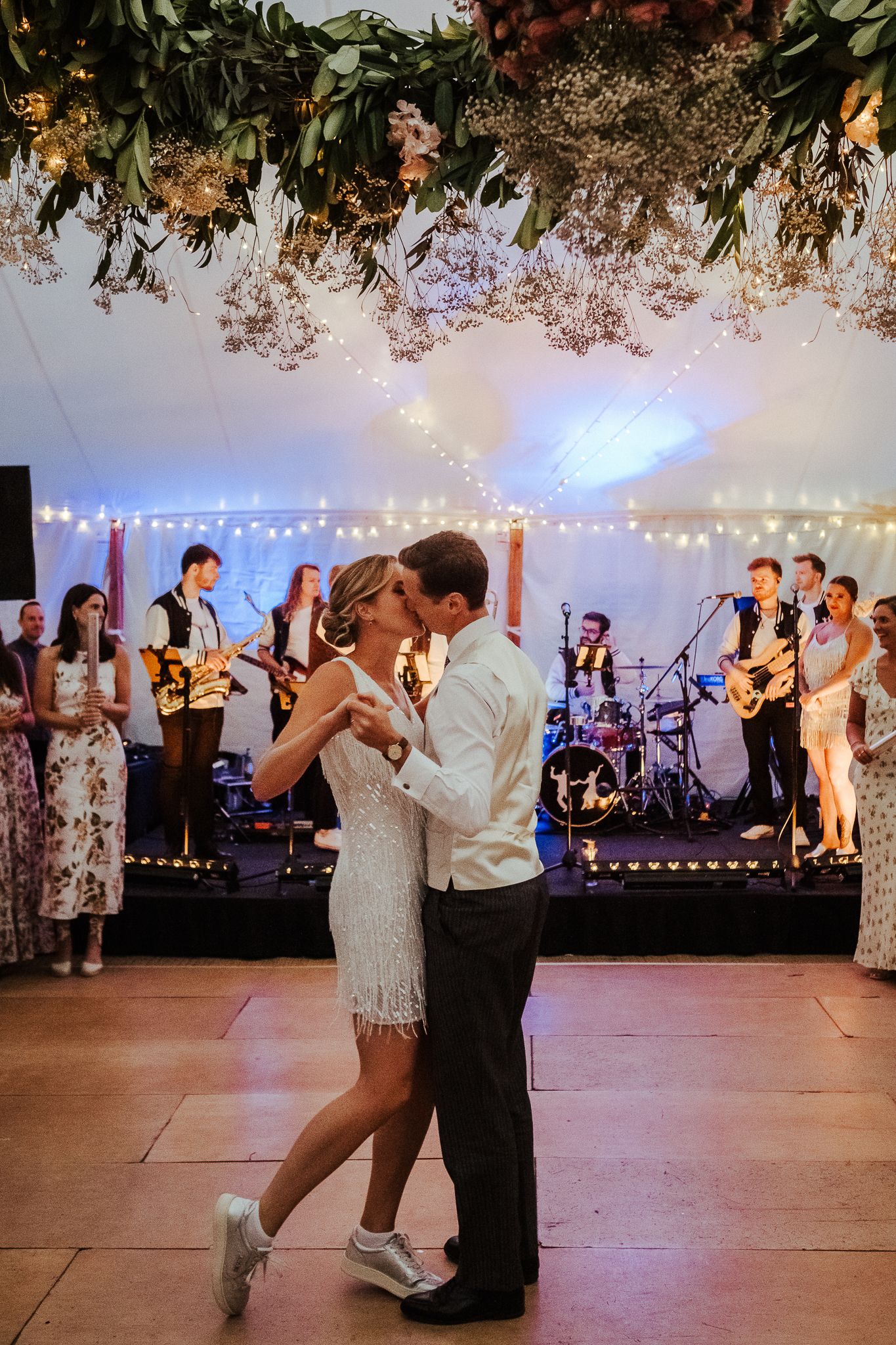 First dance at wedding reception with live band performing under hanging floral decorations and string lights.