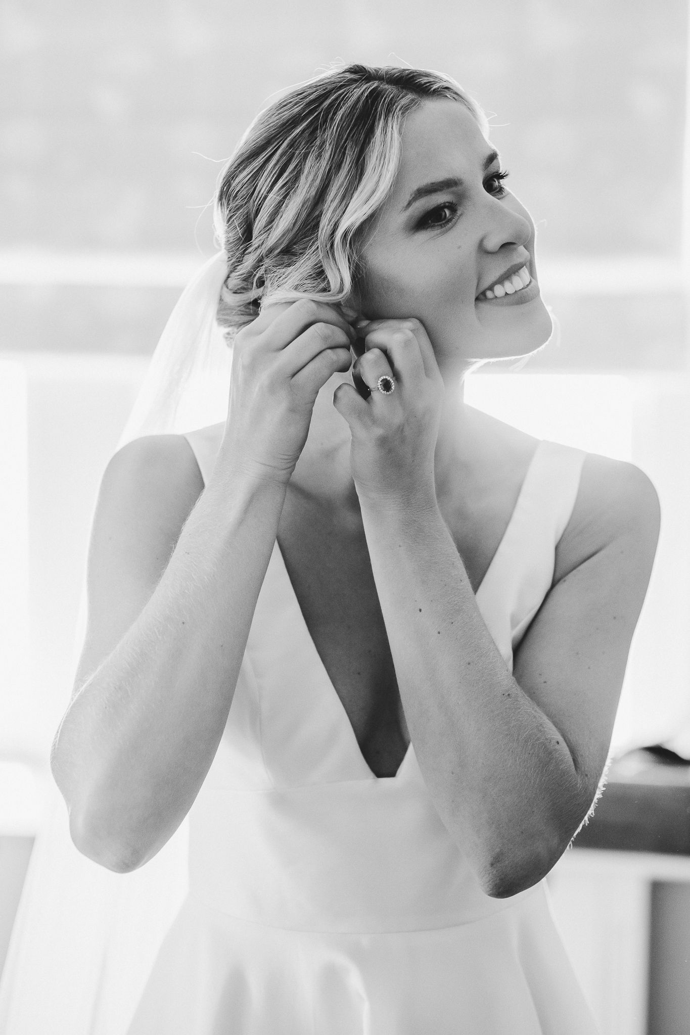 A bride in a sleeveless white dress putting on earrings while getting ready for her wedding.