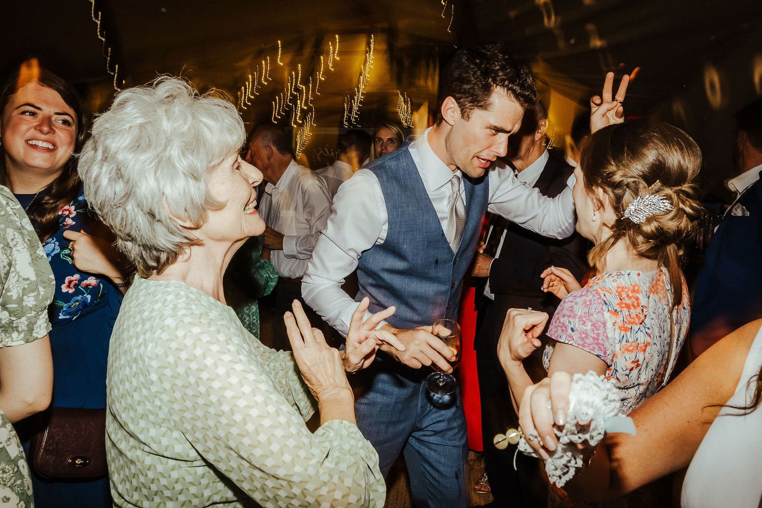 Wedding guests of all ages dancing together at a reception under decorative lighting.