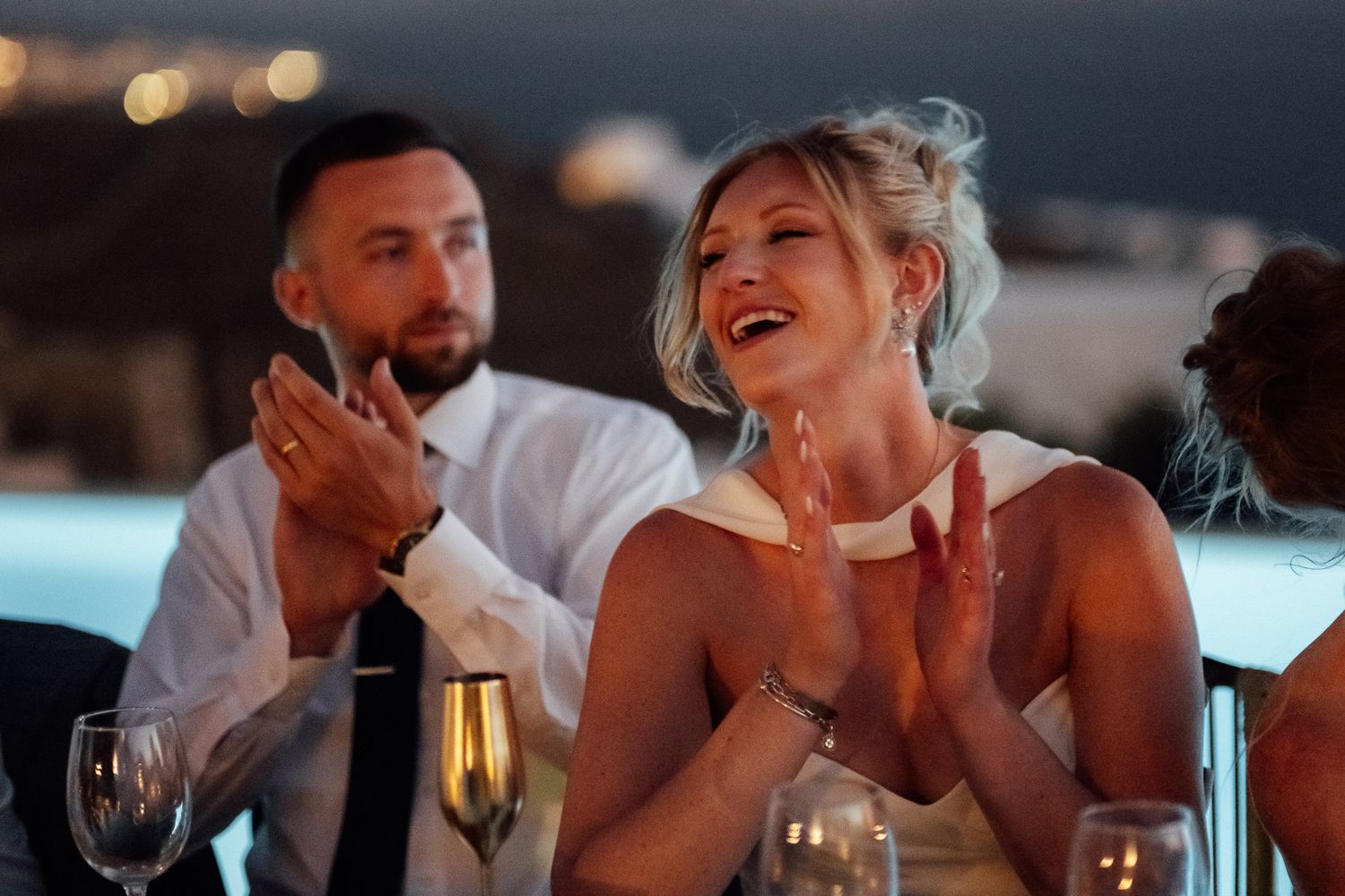 Bride and groom laughing and clapping at outdoor wedding reception dinner with city lights in background.
