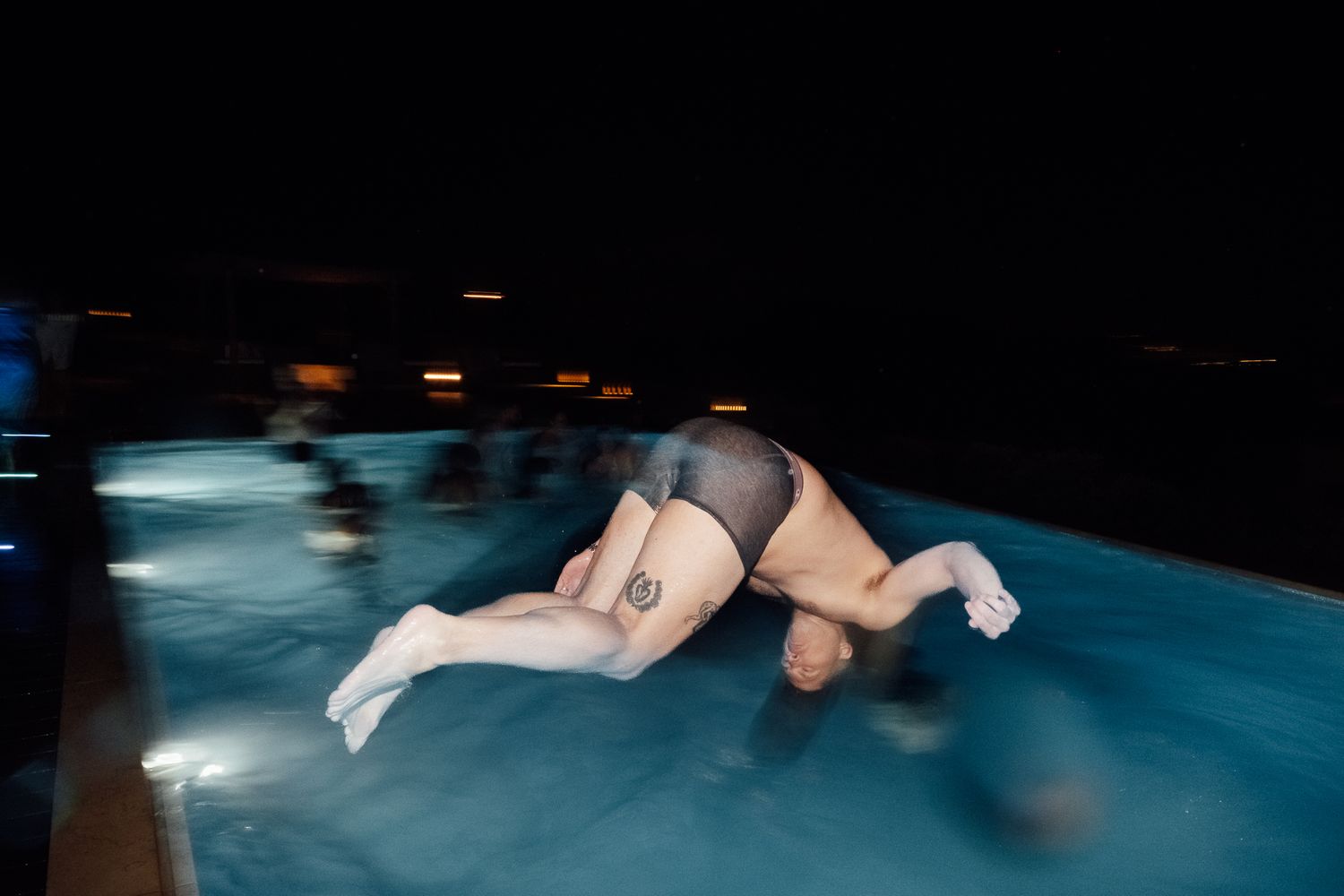 Person diving into an illuminated pool at night with city lights visible in the background.