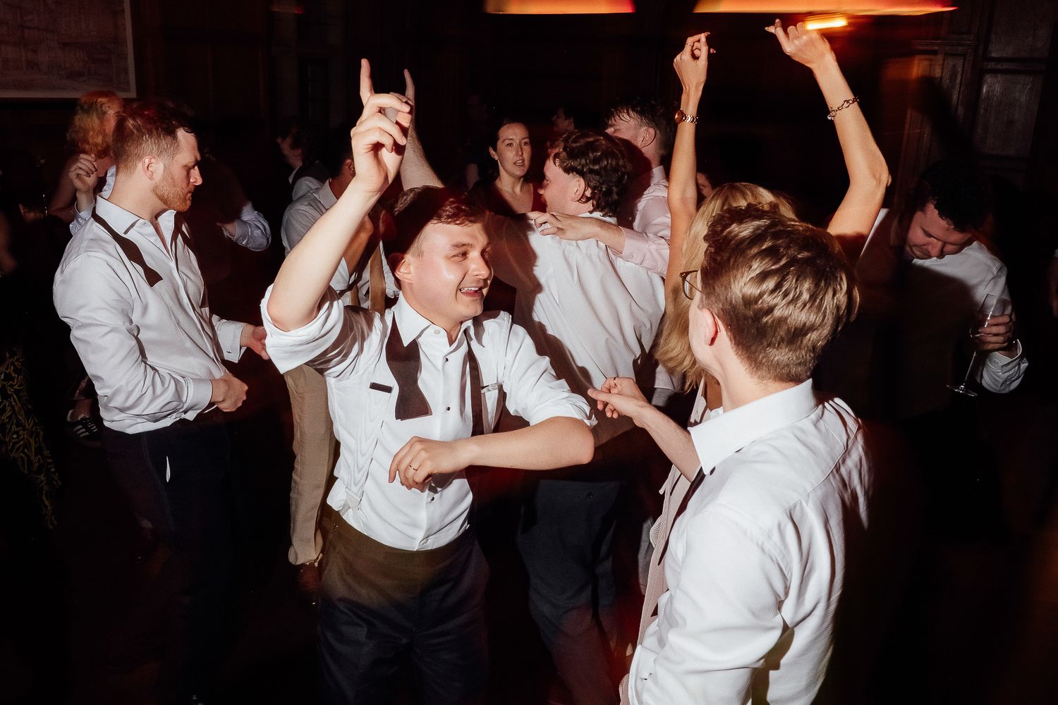 Group of young men dancing energetically at a party, wearing white shirts and suspenders in a dimly lit venue.