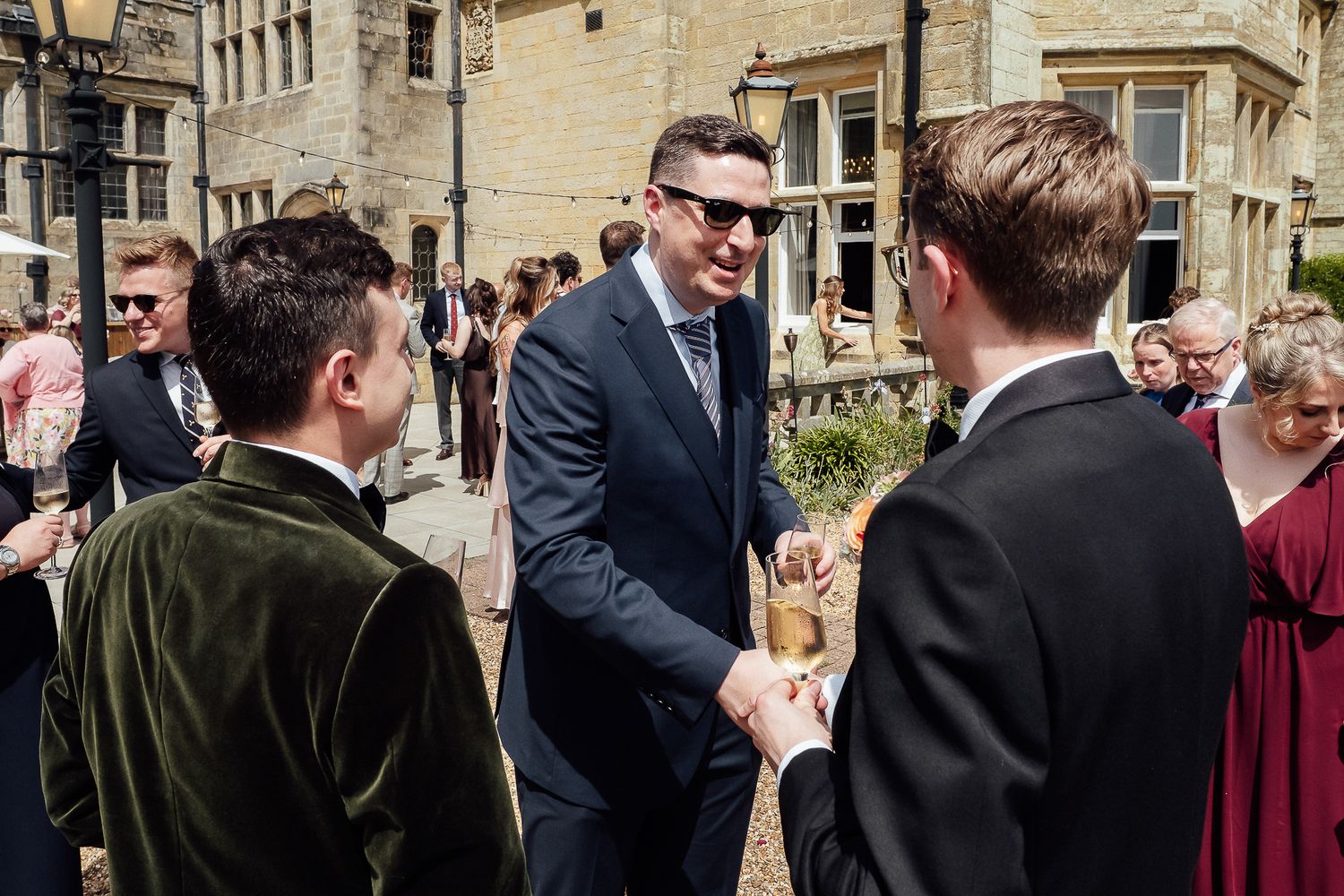 Guests in formal attire socializing outdoors at a wedding reception near a stone building on a sunny day.