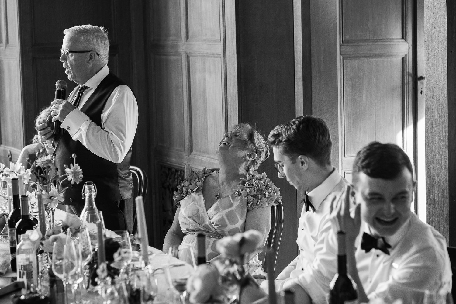 Black and white wedding reception photo of a man giving a toast while guests laugh and smile at a decorated table.