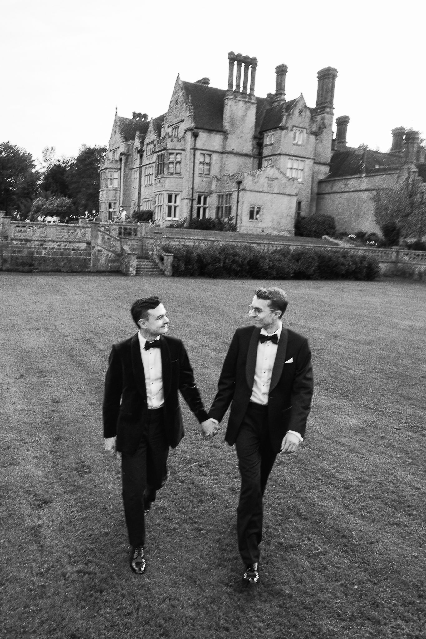 Two grooms in black tuxedos hold hands walking in front of a grand English manor in black and white.