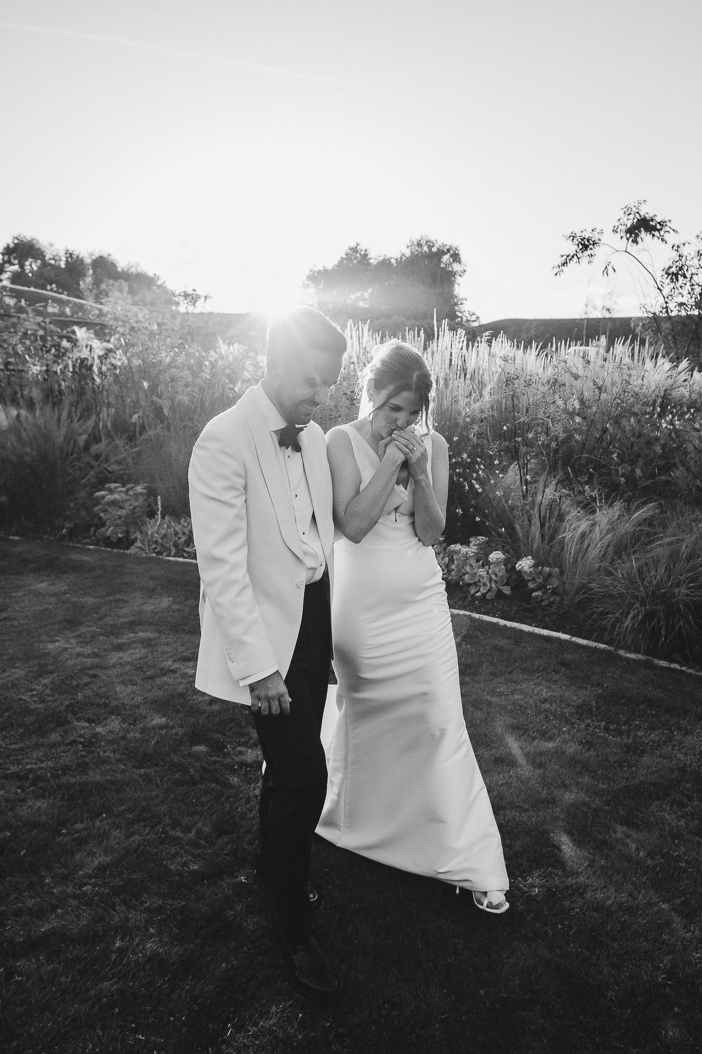 A romantic black and white wedding photo of a couple sharing a laugh together in a garden at sunset.