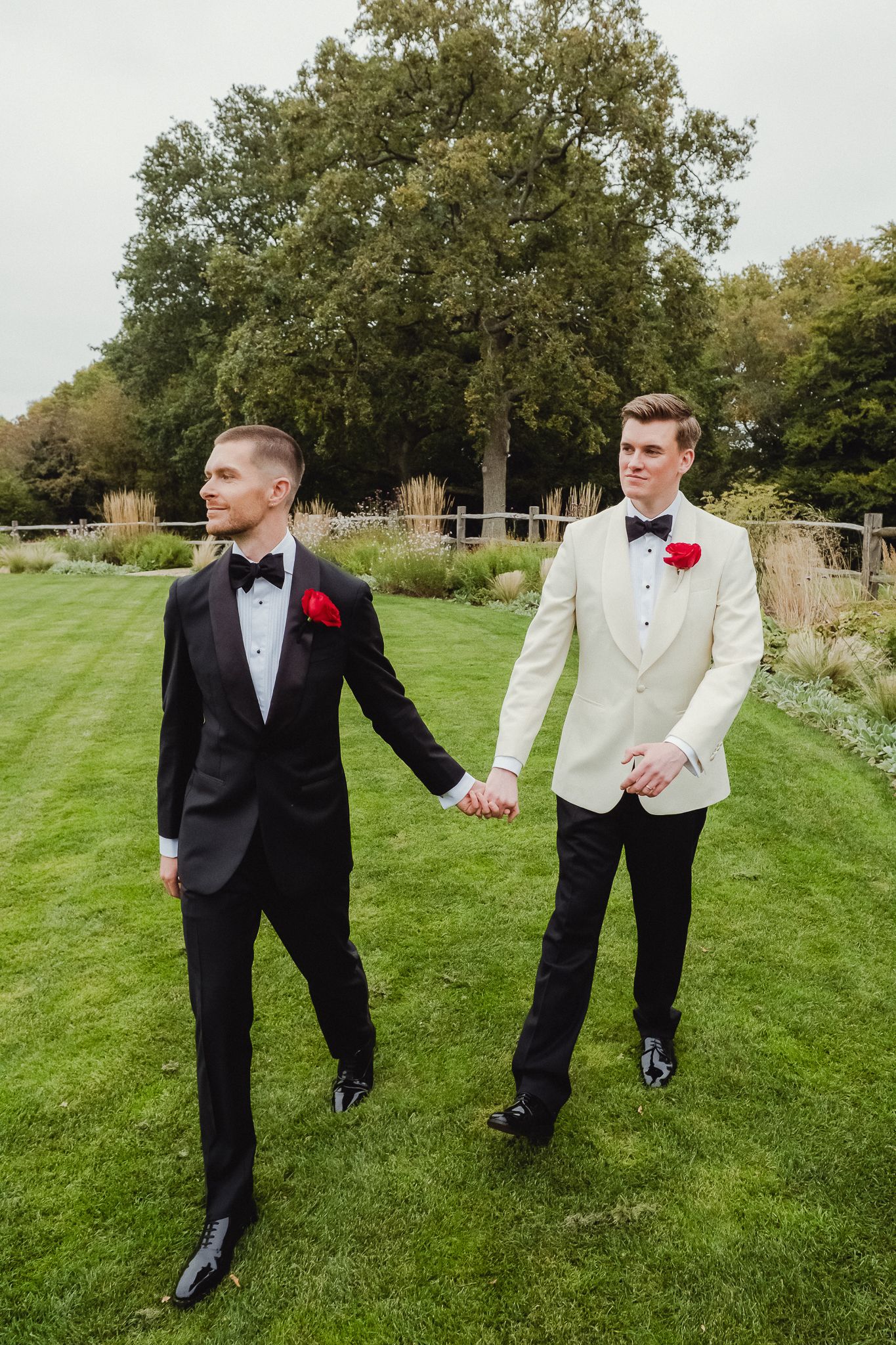 Two people in formal wedding attire walk hand in hand on a manicured lawn with trees in the background.