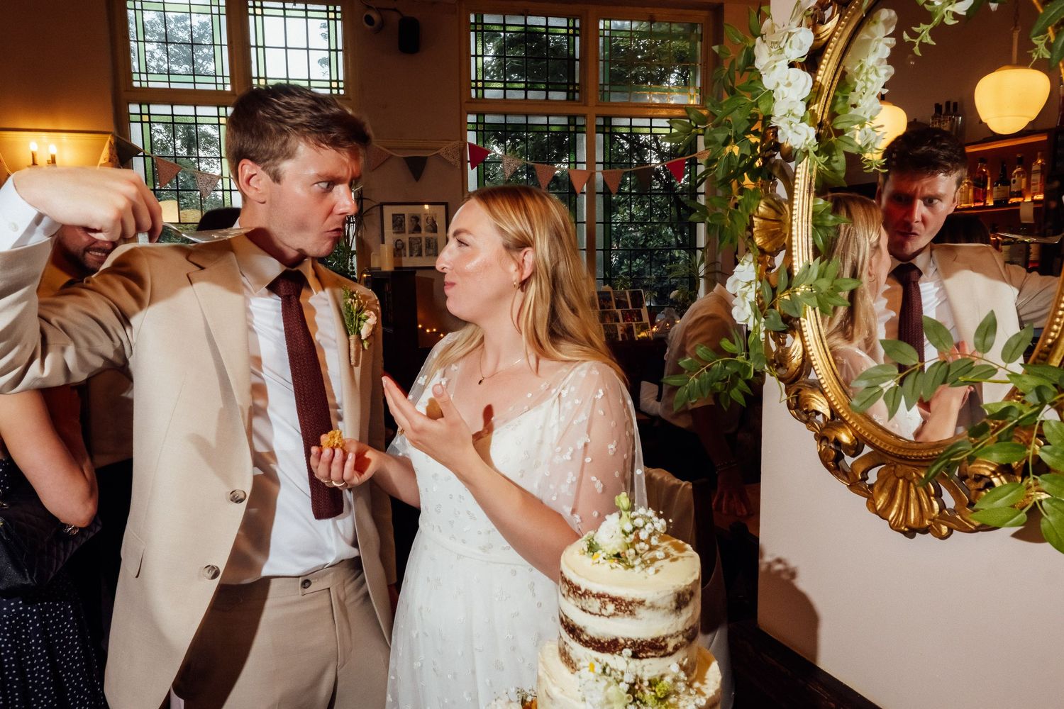 A bride in a white lace dress and a groom in a tan suit playfully feed each other wedding cake beside an ornate gold mirror.