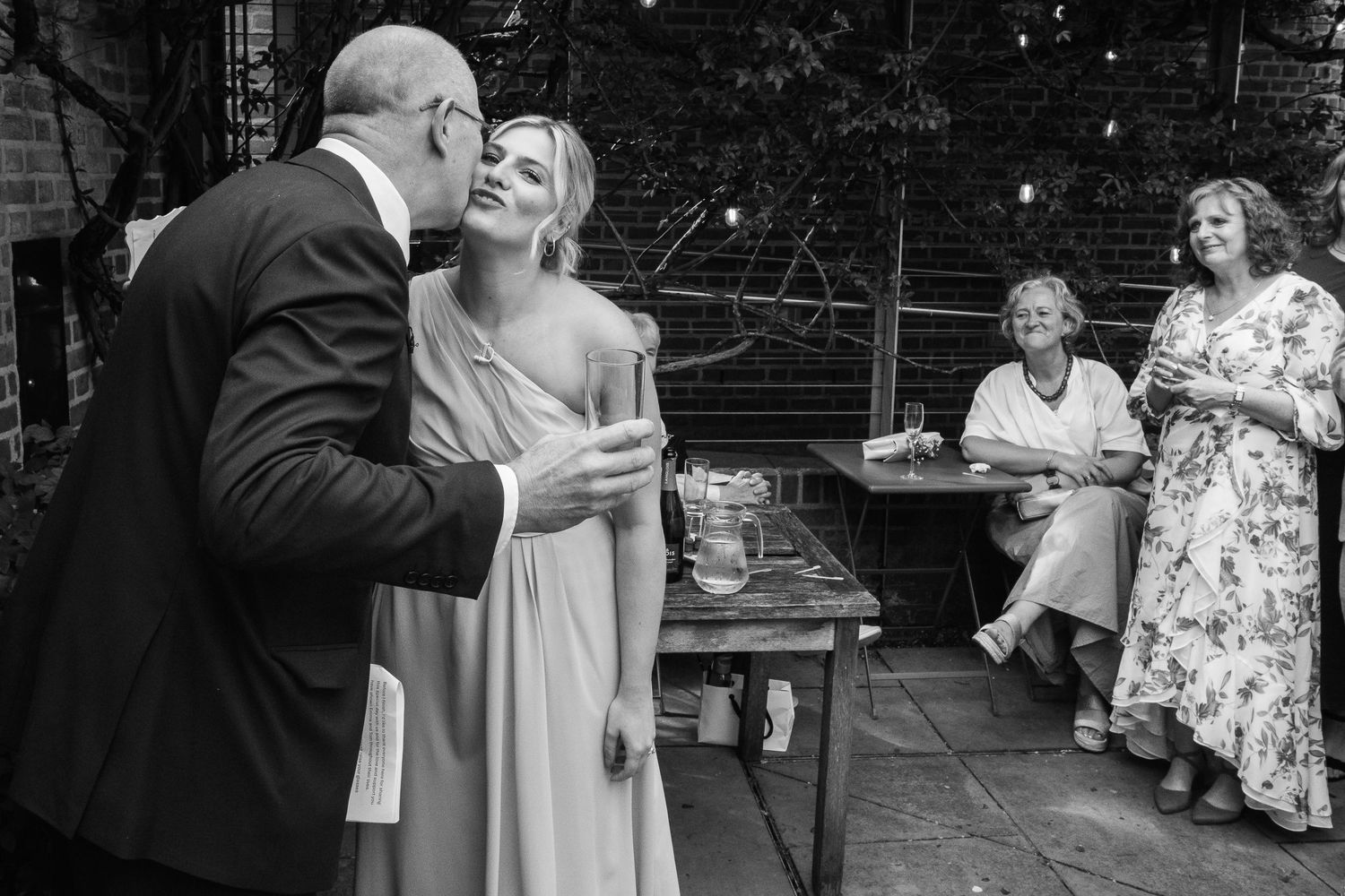 A man in a dark suit kisses a woman in a pale satin dress on the cheek while two women chat at an outdoor wedding celebration.