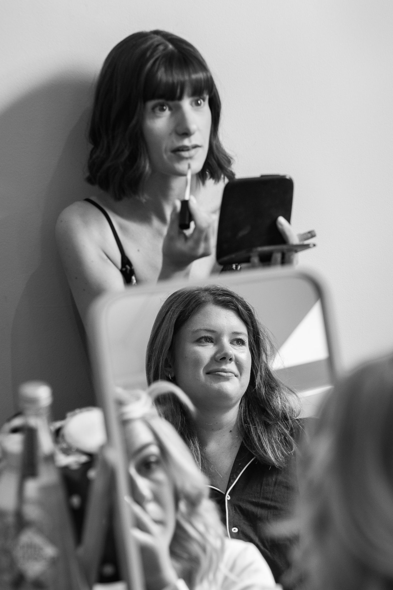 Black and white photo of a woman applying lip gloss while holding a compact mirror as another woman smiles in the foreground.