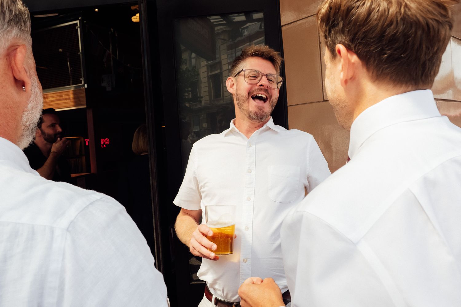 A smiling bearded man in glasses and a white shirt laughs while holding a glass of beer and chatting with two men outside a restaurant.