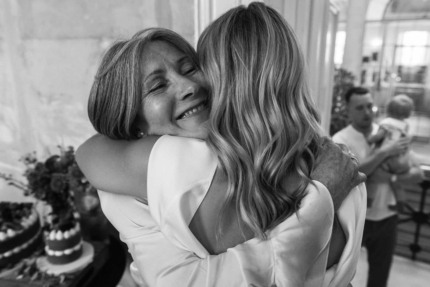 Two women share a warm, emotional hug at a celebration, with a smiling woman's face visible and a man holding a child in the background.