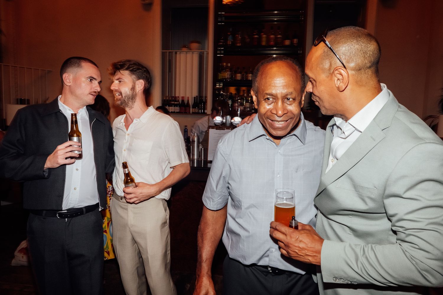 Four men laugh and chat while holding drinks at a lively indoor social gathering with a bar and shelves of bottles in the background.