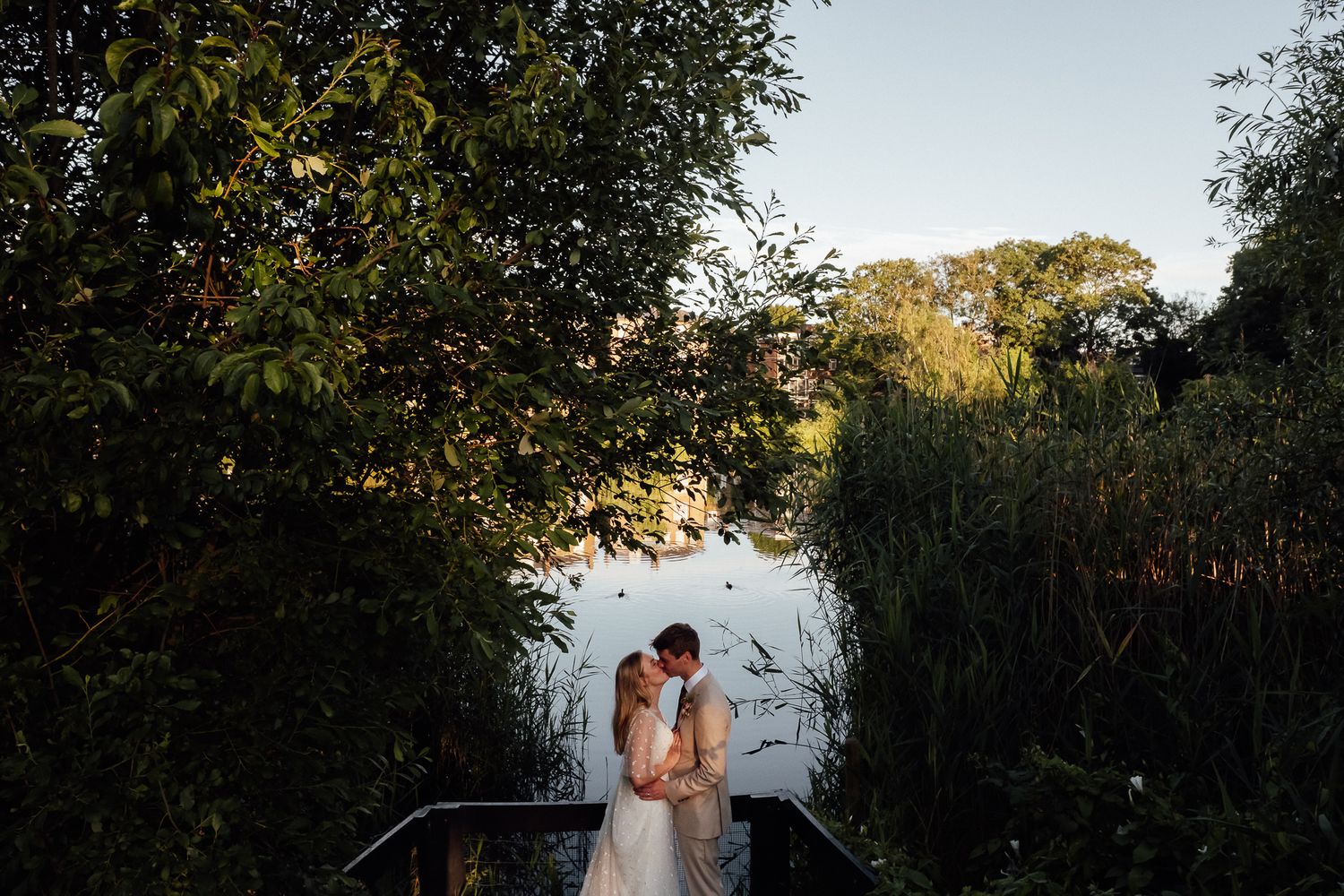 Bride and groom sharing a romantic kiss on a wooden bridge surrounded by lush greenery and a tranquil lake at golden hour.