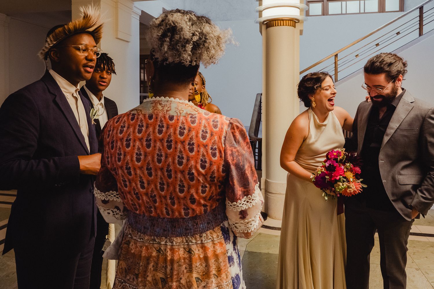 Wedding guests gather on a sunlit porch, with a person in a champagne-colored dress holding a vibrant pink bouquet.