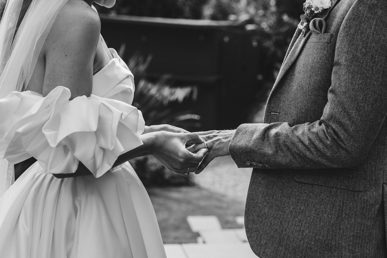 Black and white close-up of hands exchanging wedding rings during an outdoor ceremony.