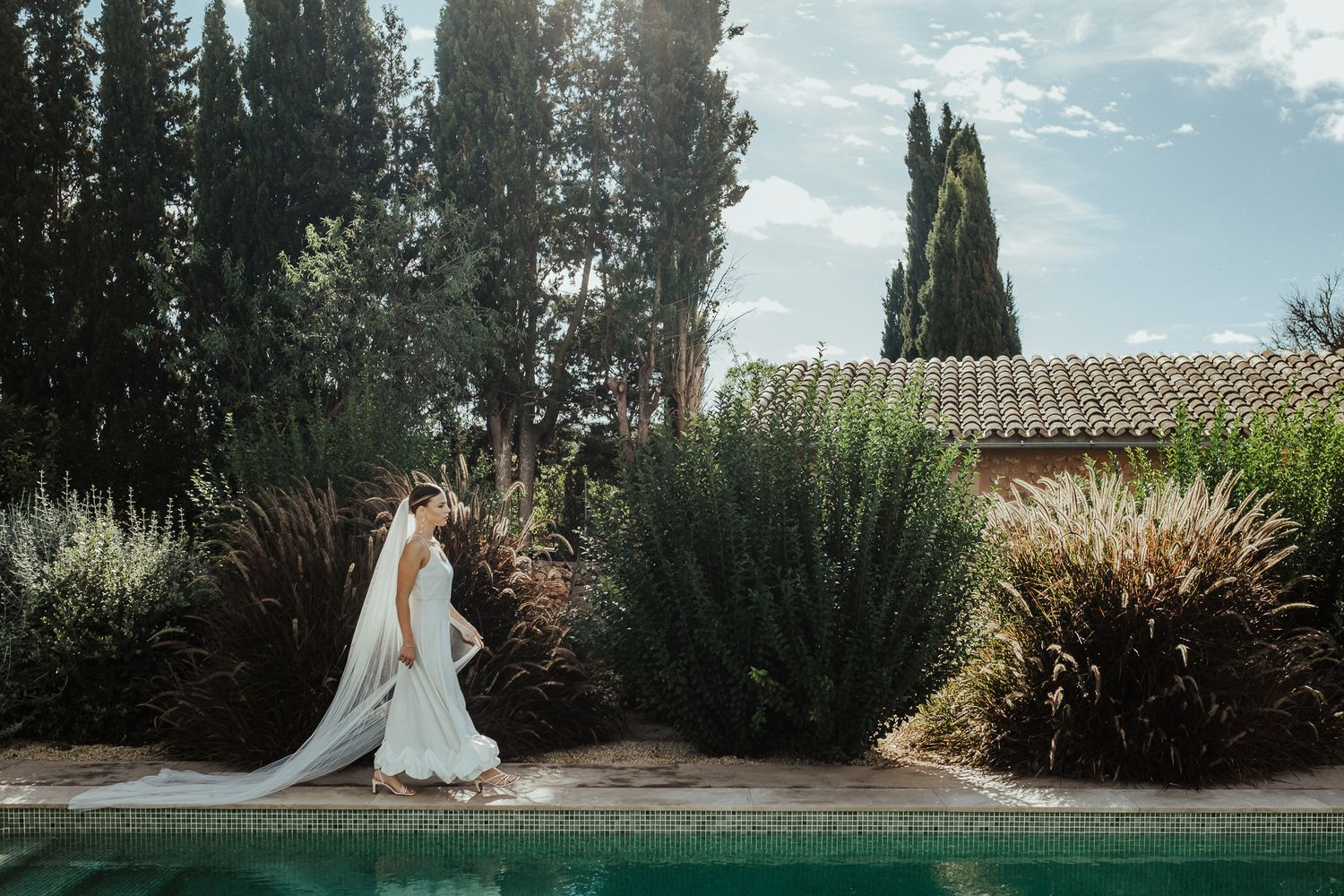 A bride in a flowing white gown poses in a serene garden setting with cypress trees and ornate architecture.