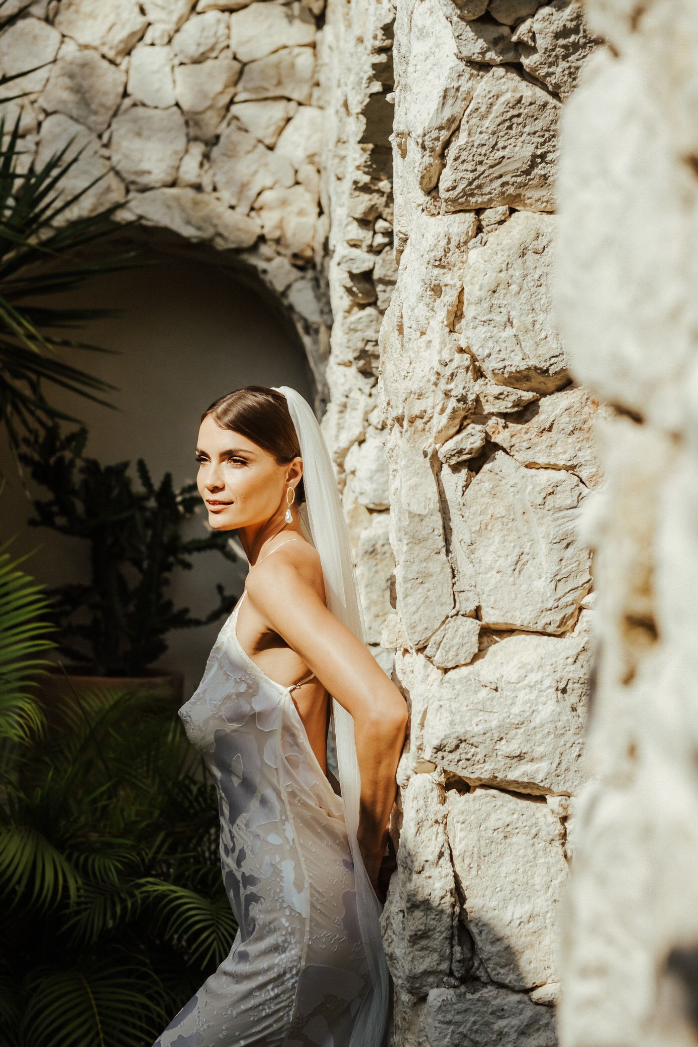 Elegant bride in a flowing white dress poses against a rustic stone wall with soft natural lighting and tropical foliage.