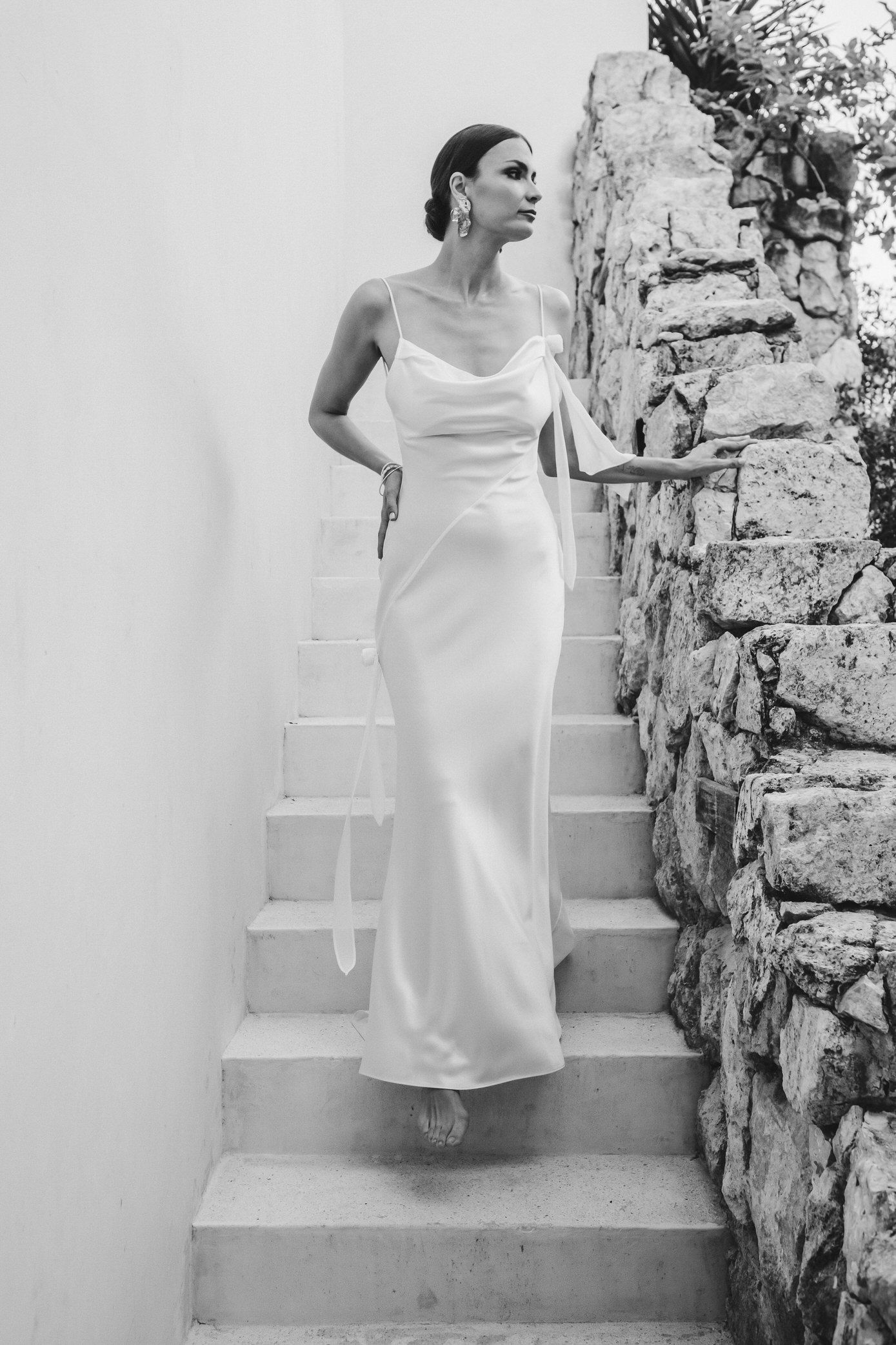 A person in a white dress stands on stone steps against a rustic wall in a black and white photograph.