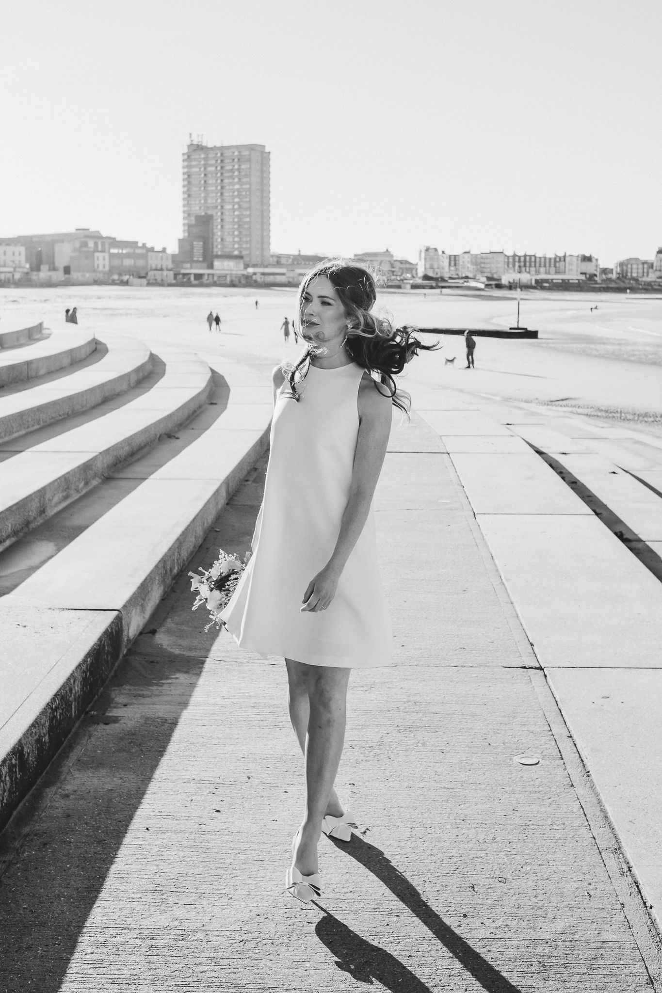 A figure in a white dress stands on a concrete walkway near the ocean with city buildings visible in the background.