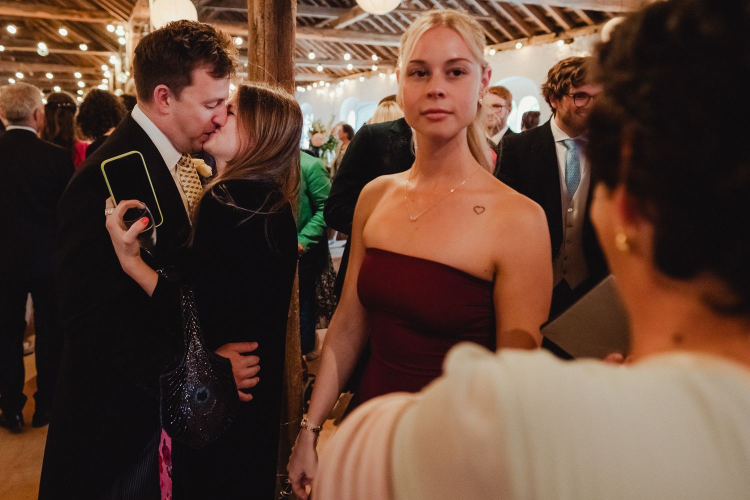 Groups of wedding guests dance and socialize on a dance floor under rustic wooden beams and warm lighting.
