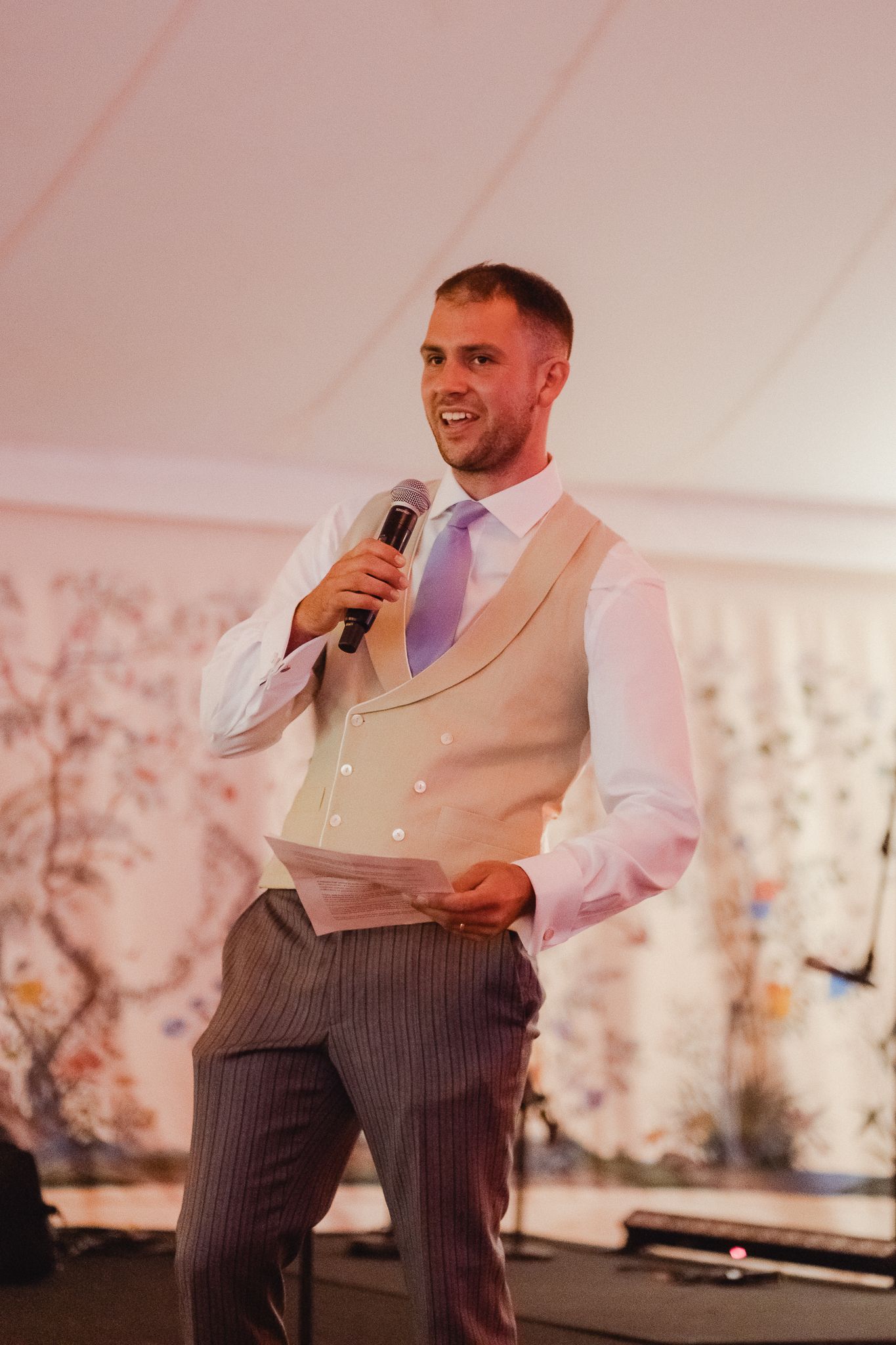 Someone gives a speech at a wedding reception wearing a beige vest, white shirt, purple tie and pinstripe pants.