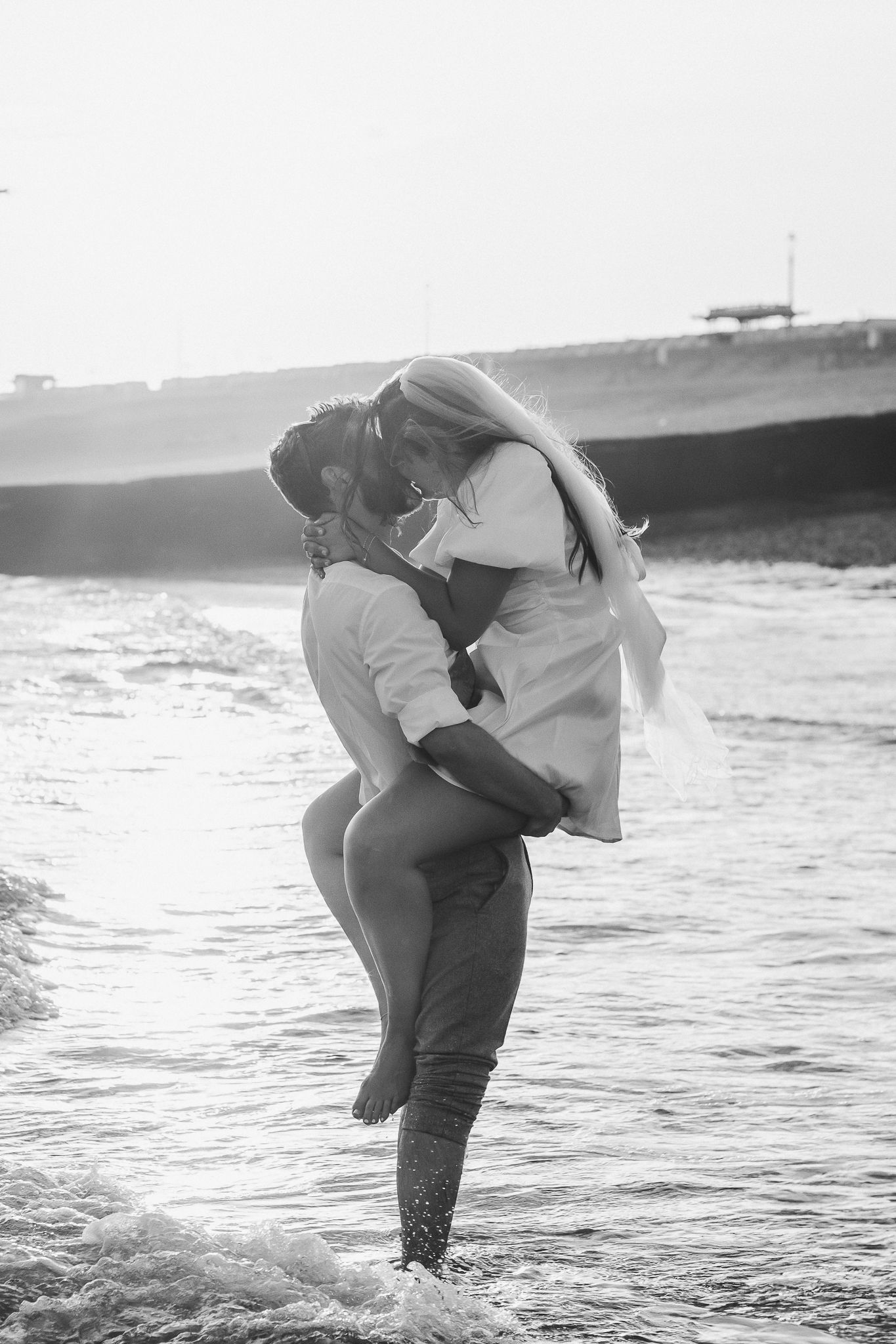 Romantic couple embraces in shallow ocean waves at sunset in black and white photo series.