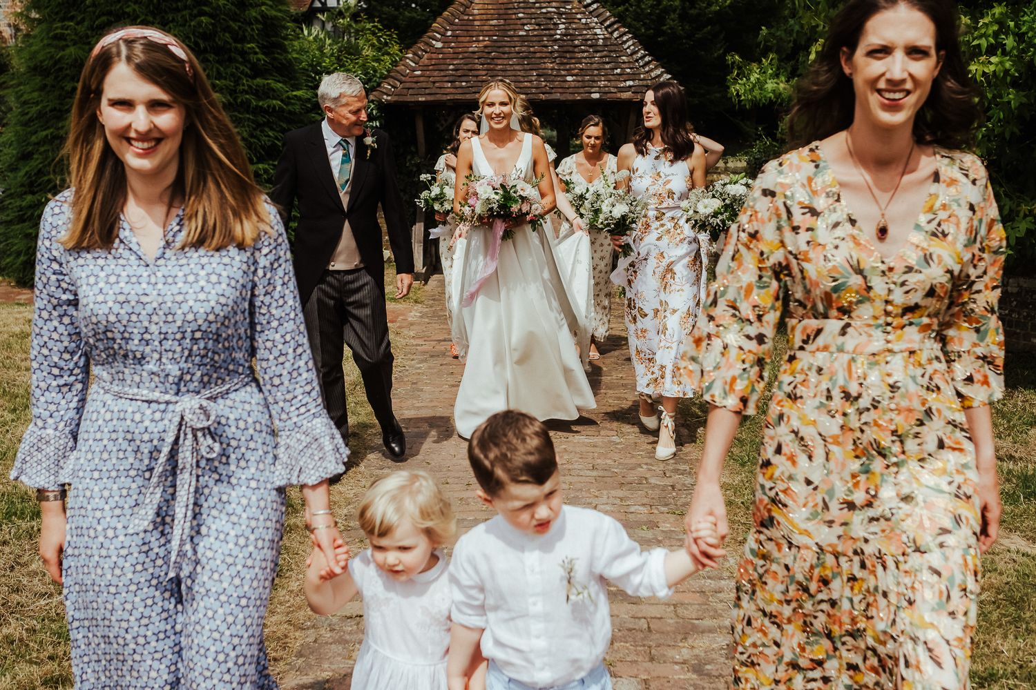 Wedding party members walk along a garden path with children in floral and patterned dresses.